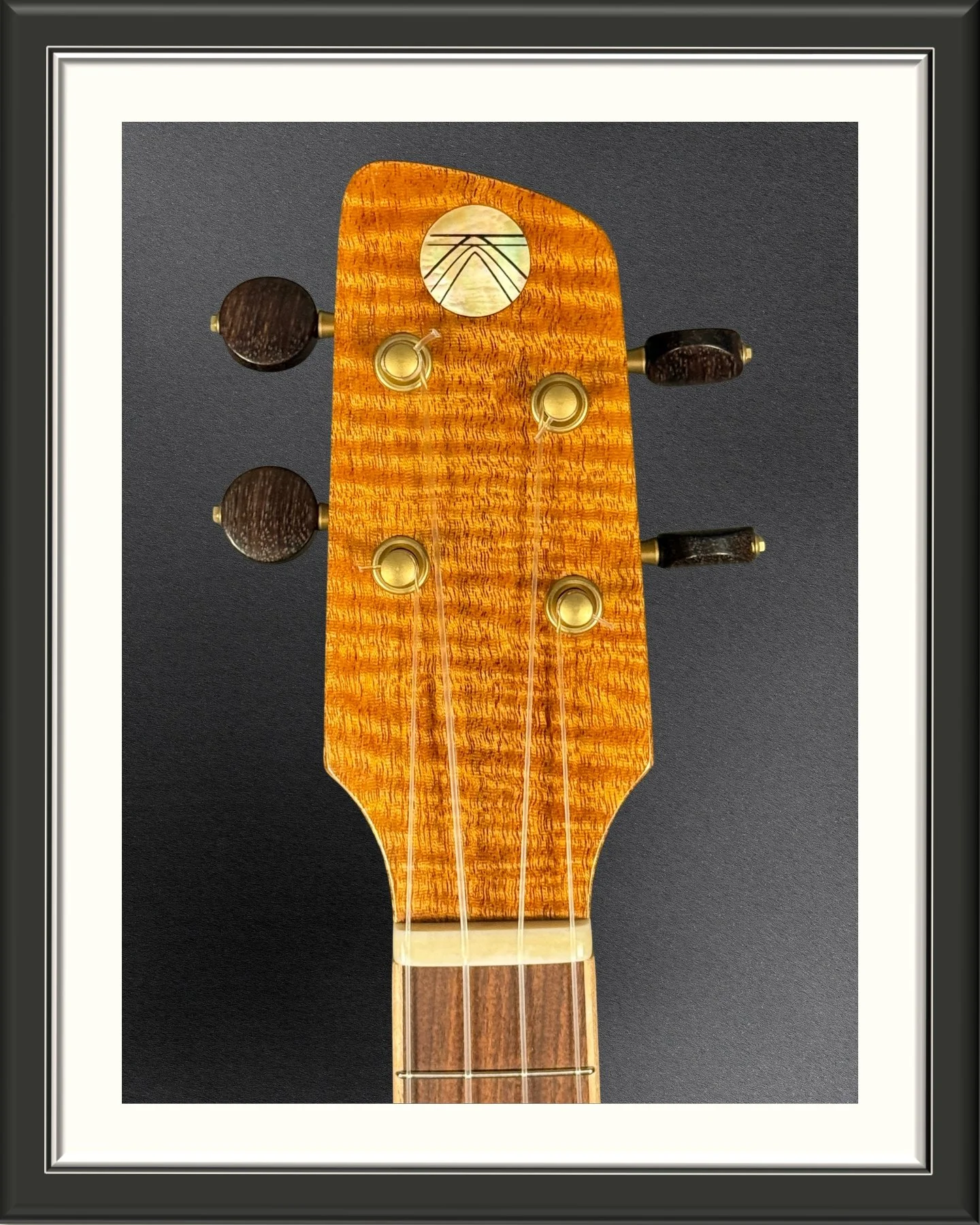 Headstock of a concert ukulele with a Curly Mahogany headplate and the MachMachines logo inlayed in mother-of-pearl