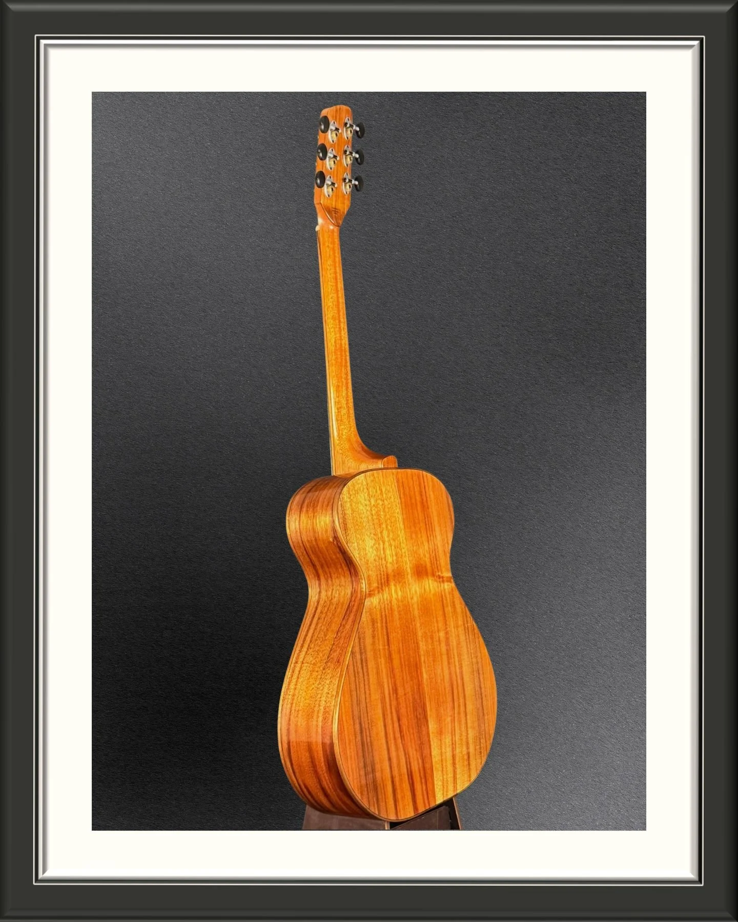 A wooden acoustic guitar with a natural finish against a dark background, framed with a white and black border.