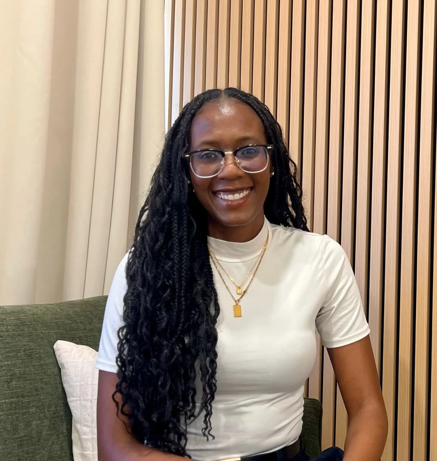 A woman with long black braided hair, glasses, gold jewelry, and a white top smiling at the camera in an indoor setting with beige curtains and wooden wall paneling behind her.