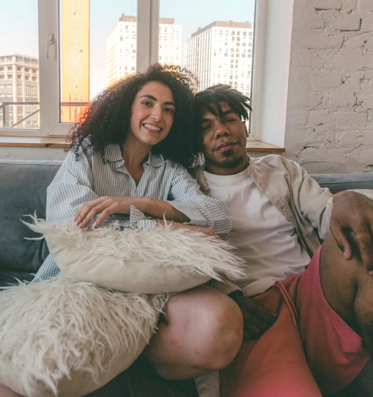 Two young adults sitting on a sofa, smiling at the camera. The woman has curly dark hair and is wearing a striped shirt. The man has dreadlocks and is wearing a beige jacket and white t-shirt. A white fluffy pillow is on the sofa in front of them. Behind them is a large window showing city buildings.