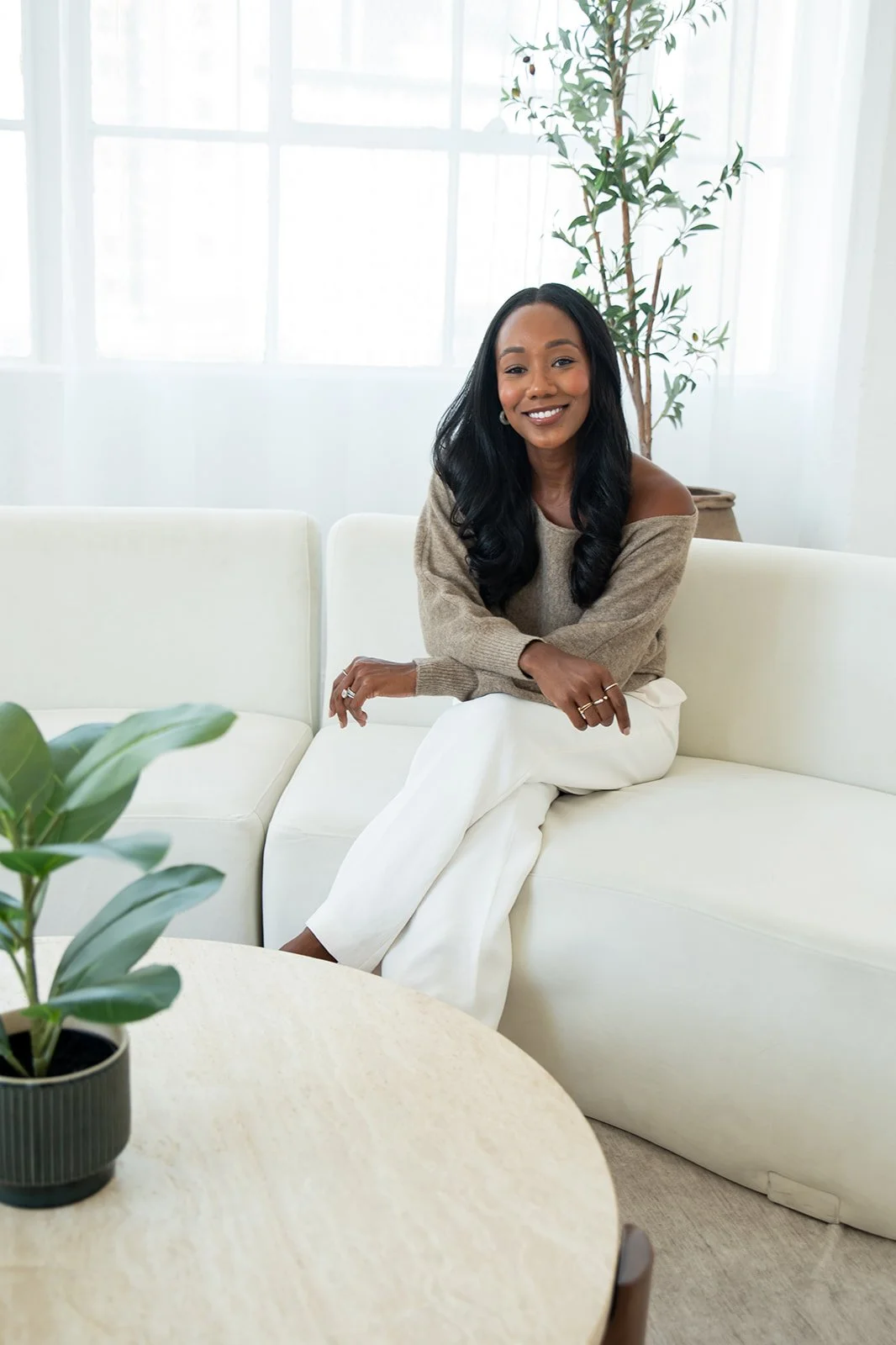 A woman with long black hair, wearing a beige sweater and white pants, sitting on a white sofa with a smile, in a bright room with large windows and a tall potted plant behind her.