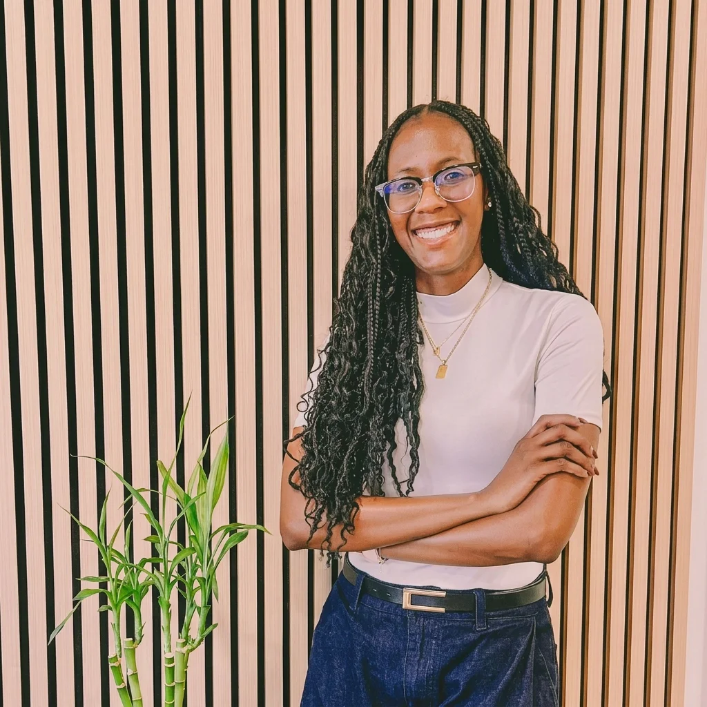 A woman with long braided hair, wearing glasses and a white top, standing with her arms crossed in front of a striped wooden wall, smiling.