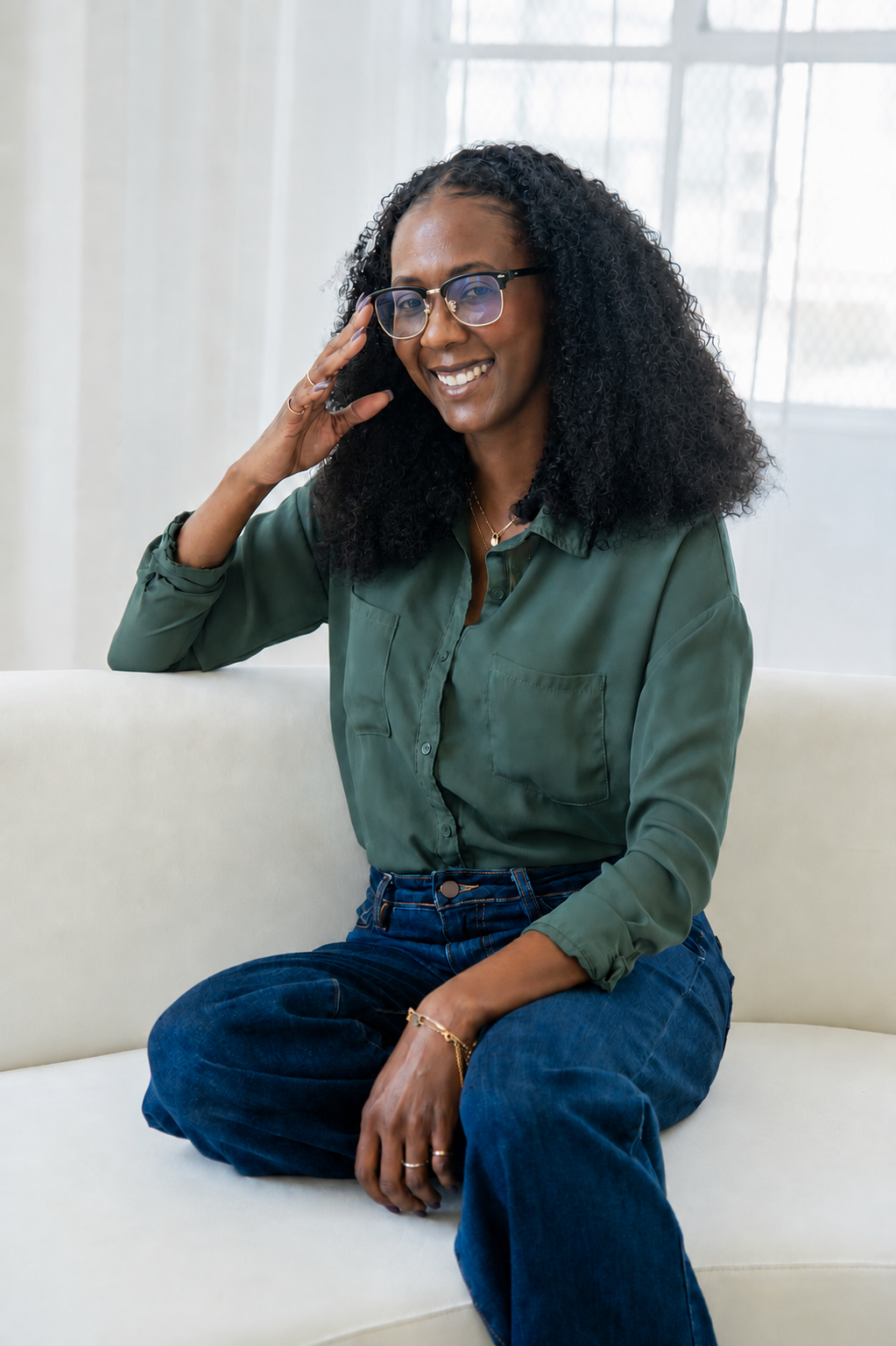 A woman with curly black hair, glasses, and a green button-up shirt sitting on a white couch, smiling and touching her glasses with her right hand indoors near a window with light curtains.