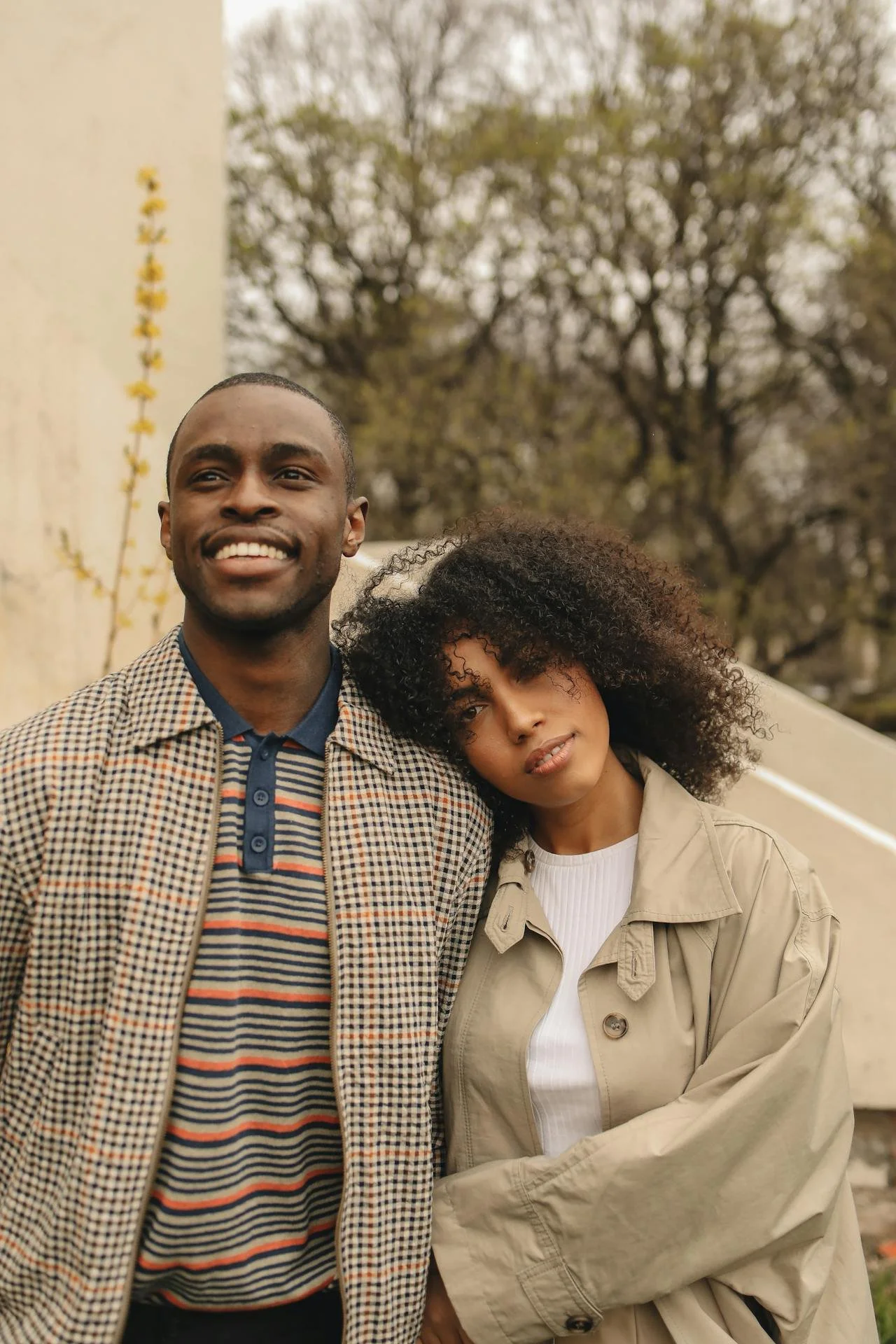 A smiling man and a woman with curly hair leaning on each other outdoors, with trees in the background.