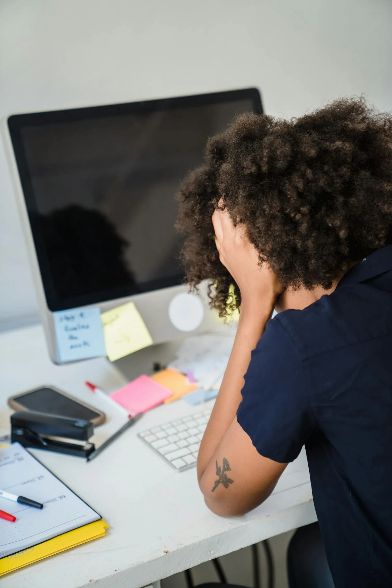 Person with curly hair sitting at a cluttered desk, holding their head in their hands in front of a computer monitor.