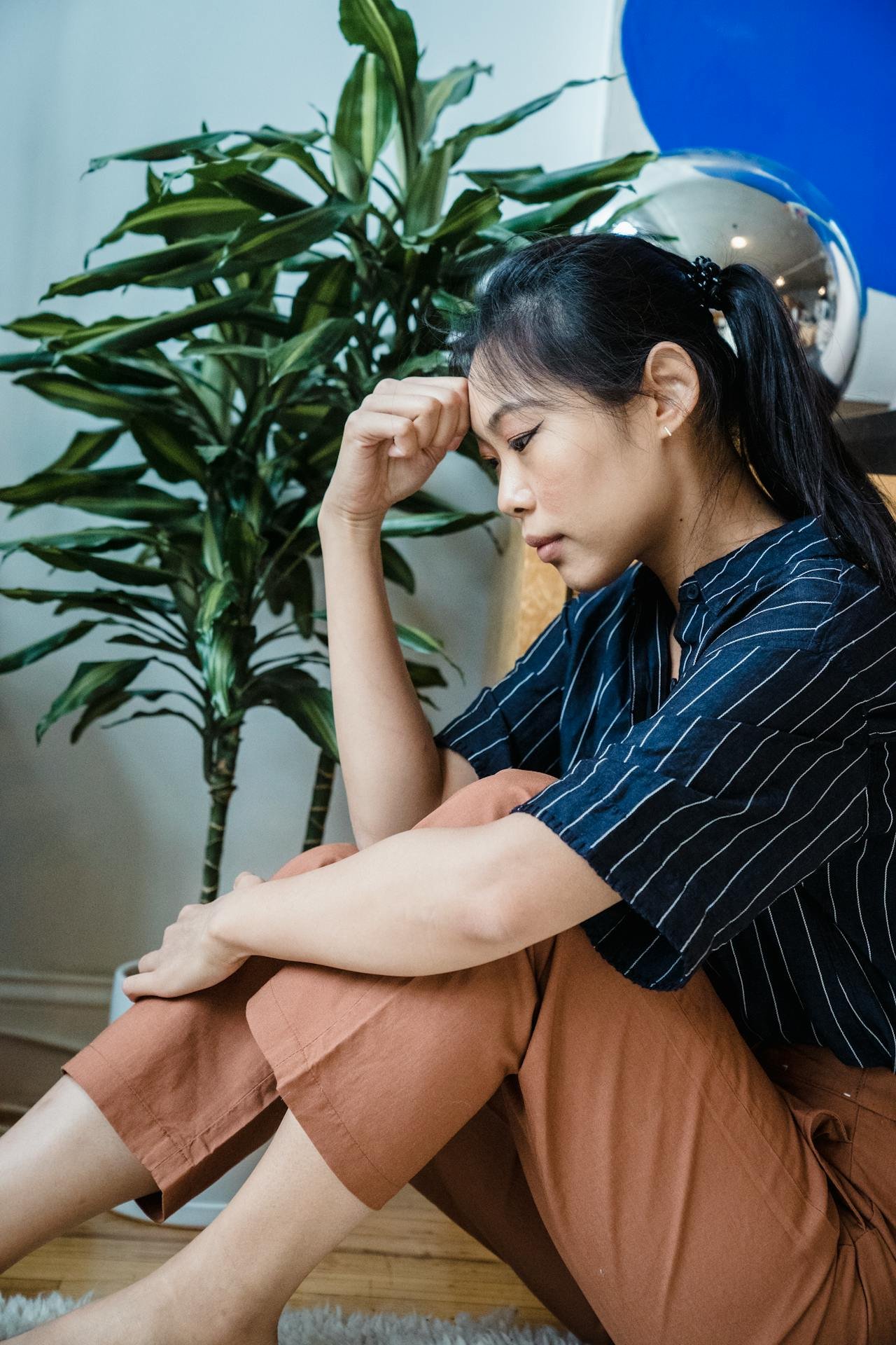 A woman sitting on the floor with her head resting on her knee, looking down with a worried or contemplative expression, in a room with a large green plant, a shiny spherical ornament, and a blue wall in the background.