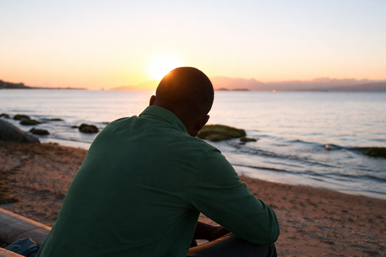 Man sitting on a beach at sunset, looking out at the water.