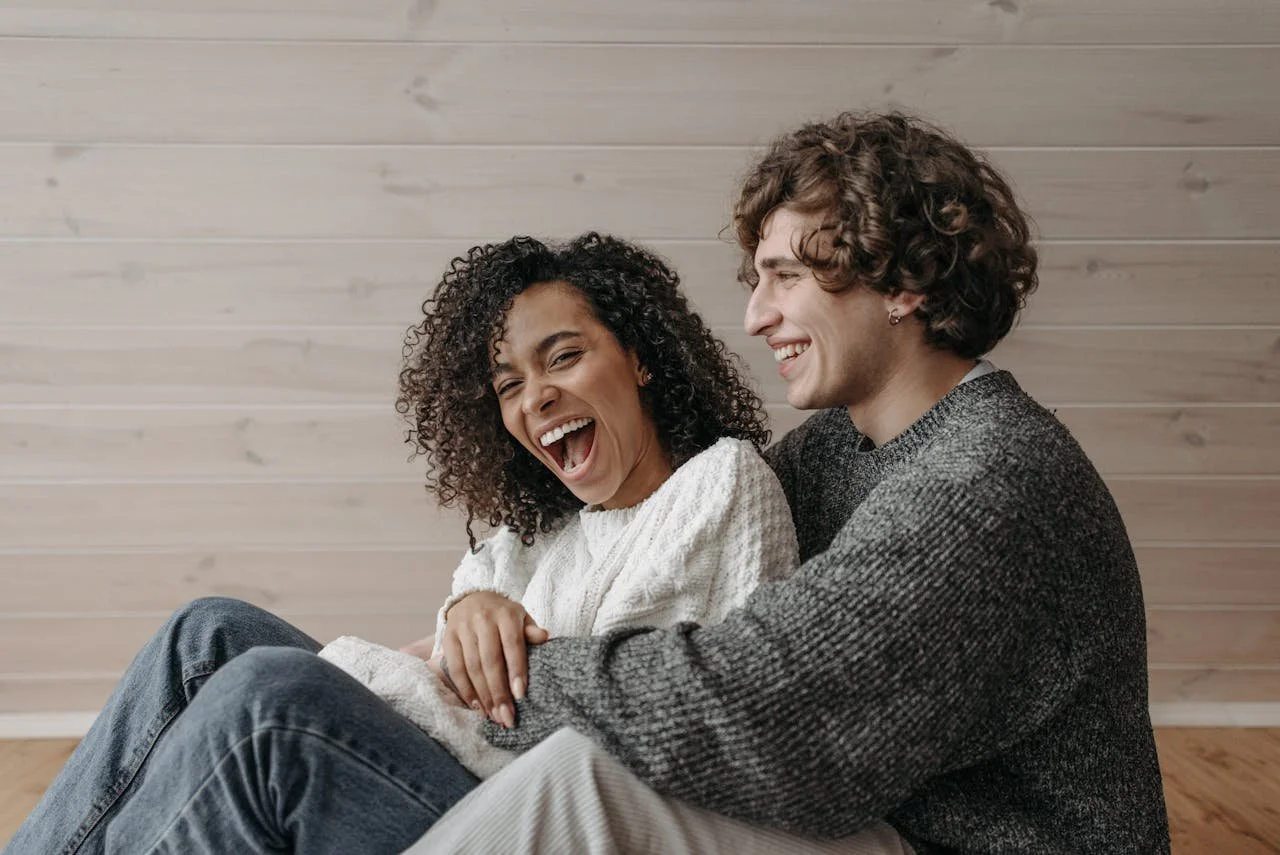 A happy couple sitting close together, laughing and smiling, indoors against a light wooden wall.