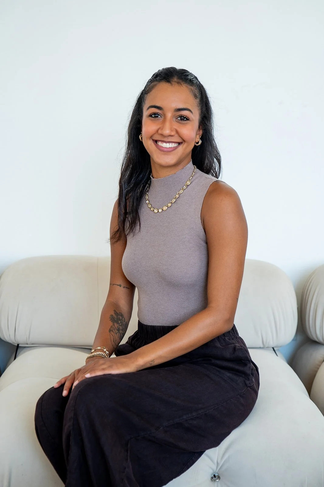 A smiling woman with dark hair, wearing a sleeveless gray top, black pants, and gold jewelry, sitting on a white sofa in a bright room with a plain white wall background.