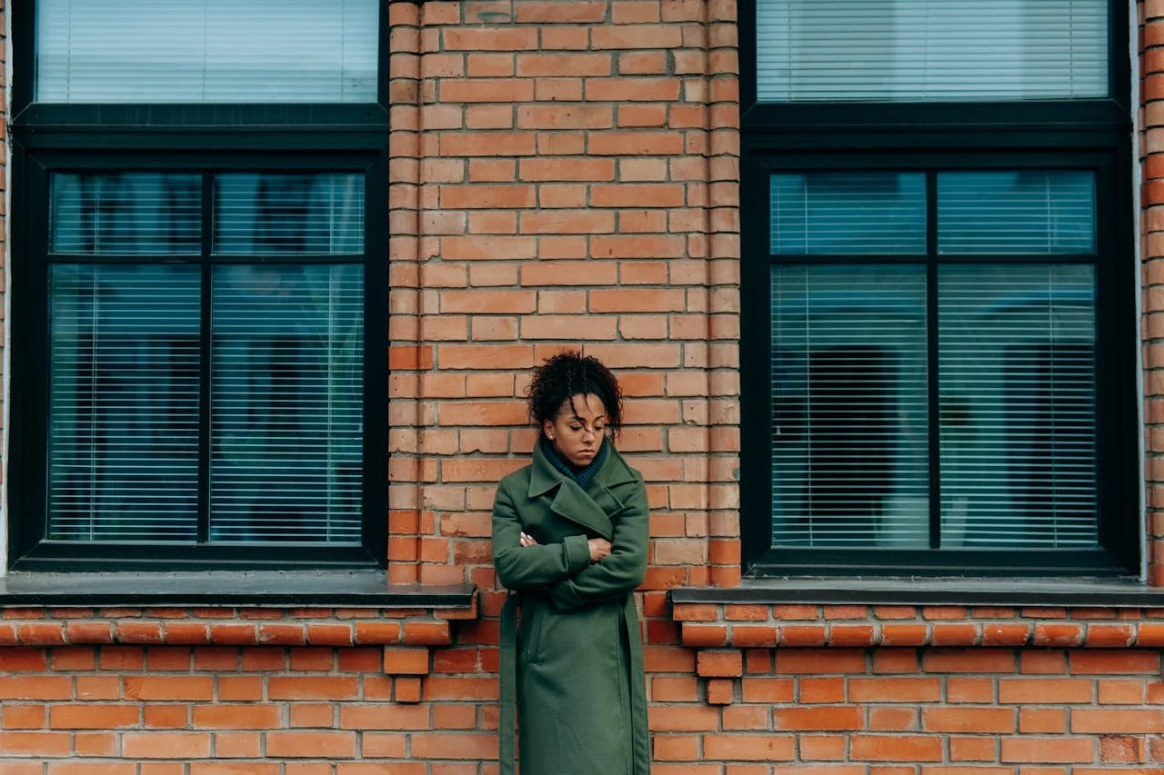 A woman with curly hair standing with arms crossed in front of a brick wall with two large windows.