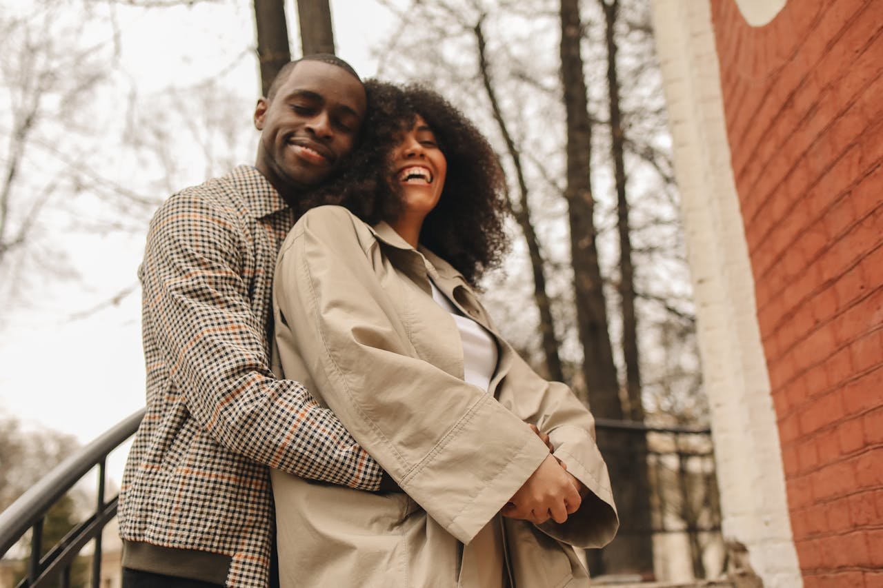 A smiling man and woman hugging outdoors near a brick wall, with trees in the background.
