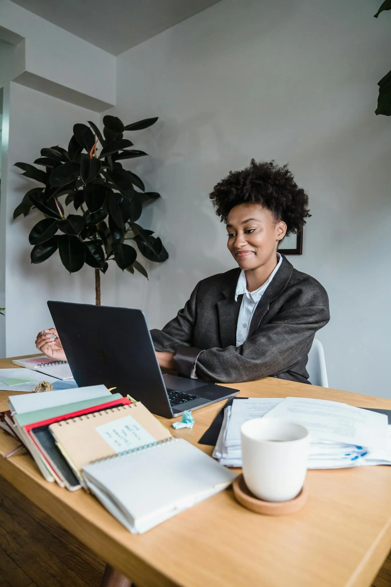 A woman in a gray blazer sitting at a cluttered wooden desk, working on a laptop with papers, notebooks, and a white mug.