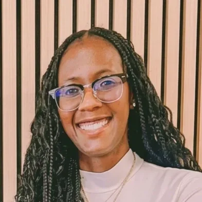 A woman with long braided hair, glasses, and a white top smiling in front of a wooden slat background.