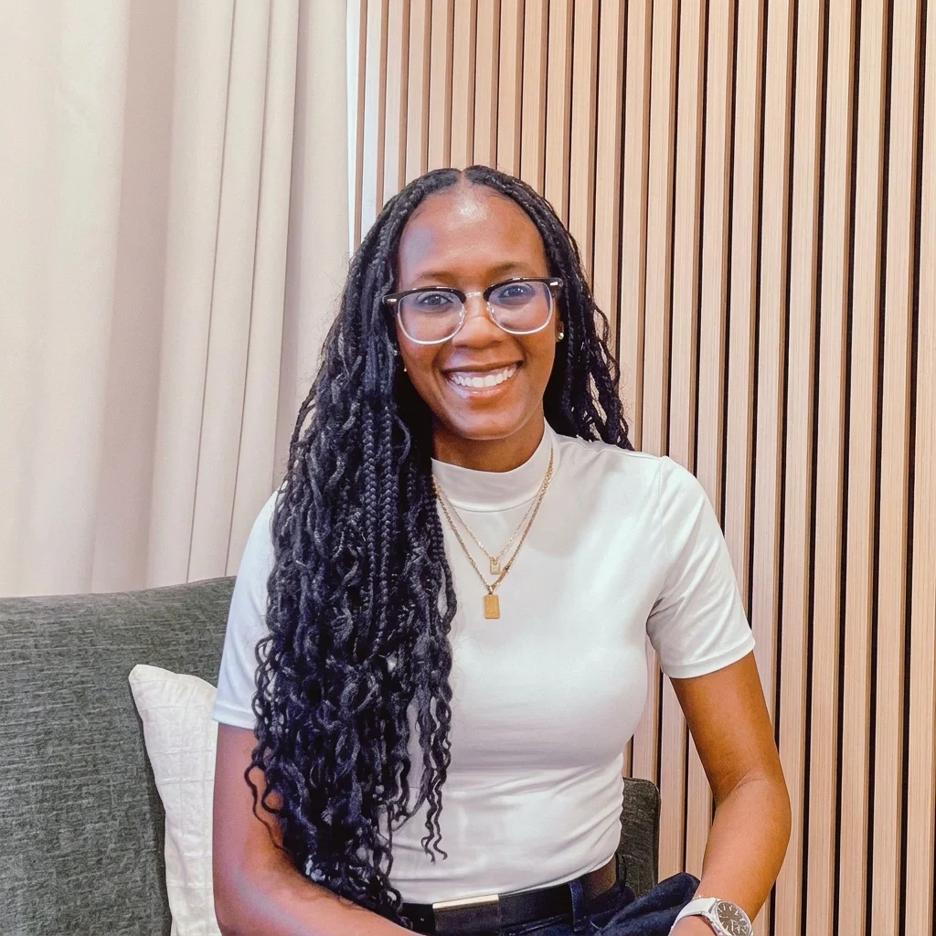 A smiling woman with long curly black hair, glasses, and a white t-shirt sitting on a gray couch in front of a wood-paneled wall.