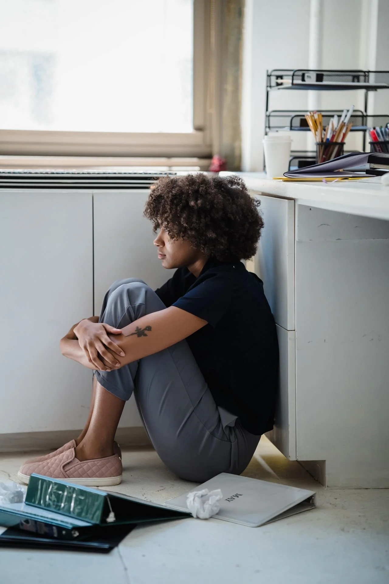 Person sitting on the floor of an office, leaning against a cabinet with an indifferent expression. Office supplies and papers are scattered around.