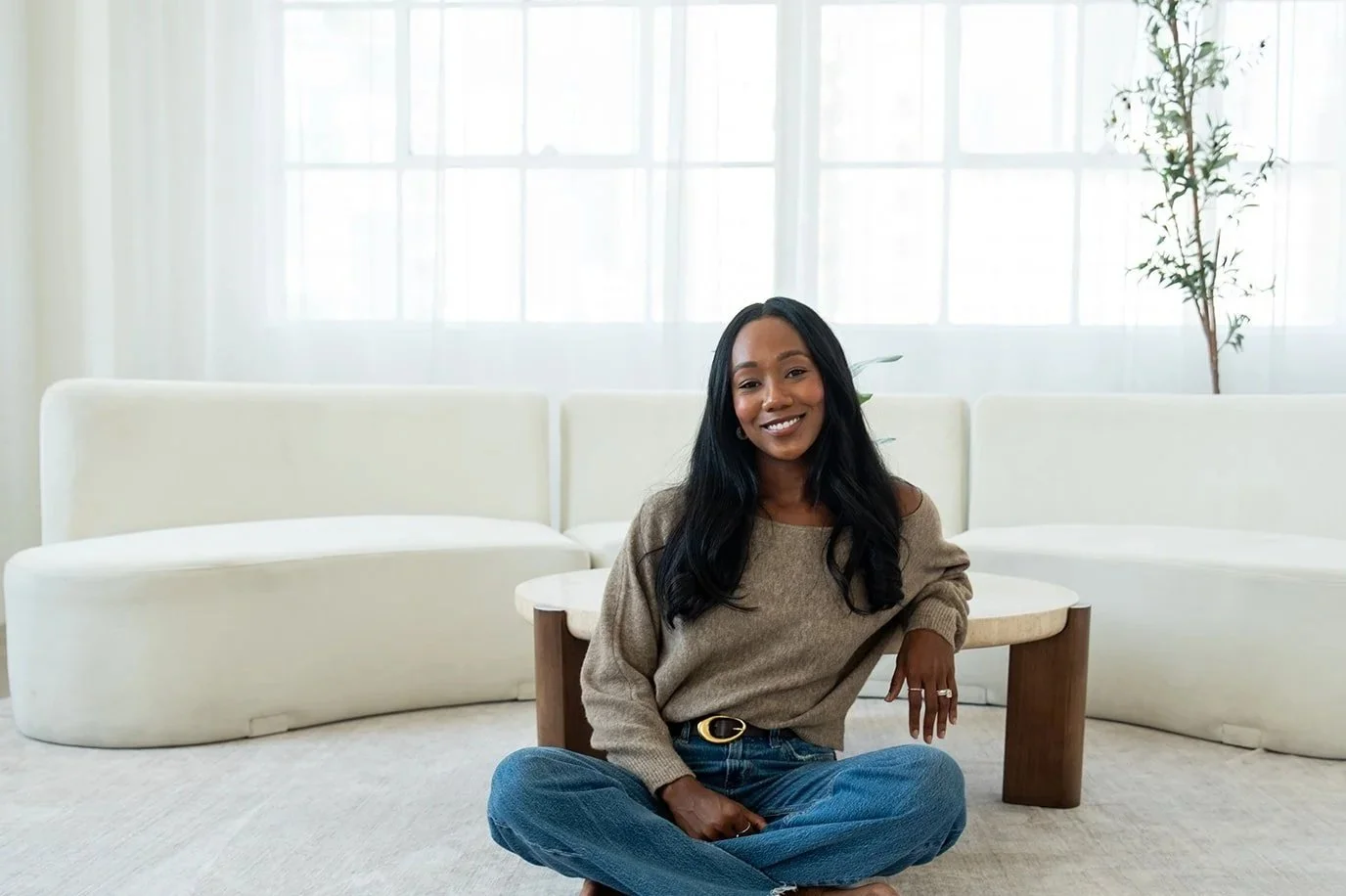 A woman with long black hair sitting cross-legged on a beige carpet in front of a modern white couch, smiling at the camera, in a bright room with large windows and a tall plant in the background.
