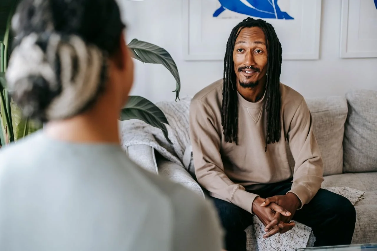 A man with long dreadlocks and a beard is sitting on a beige couch, talking to a woman whose back is to the camera. They are in a living room with framed art on the wall and a large plant nearby.