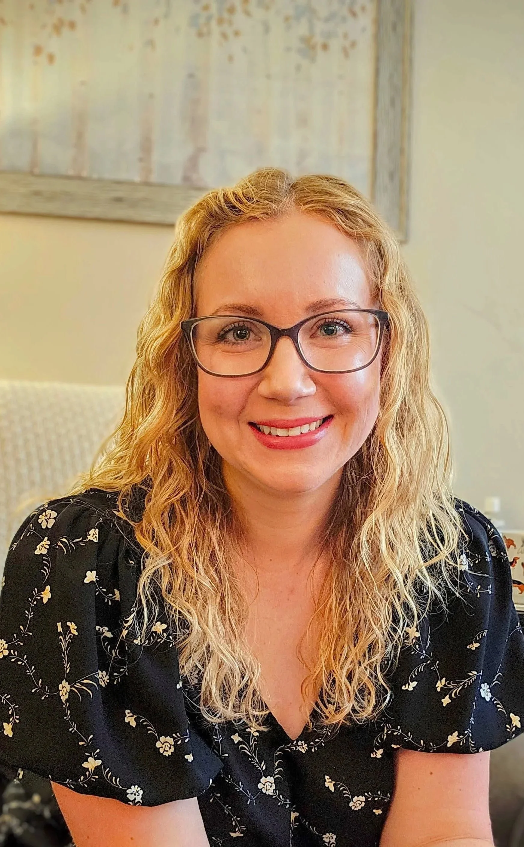 Portrait of Kristen Callaway, a woman with curly blonde hair wearing glasses and a black floral shirt, smiling indoors against a neutral background.