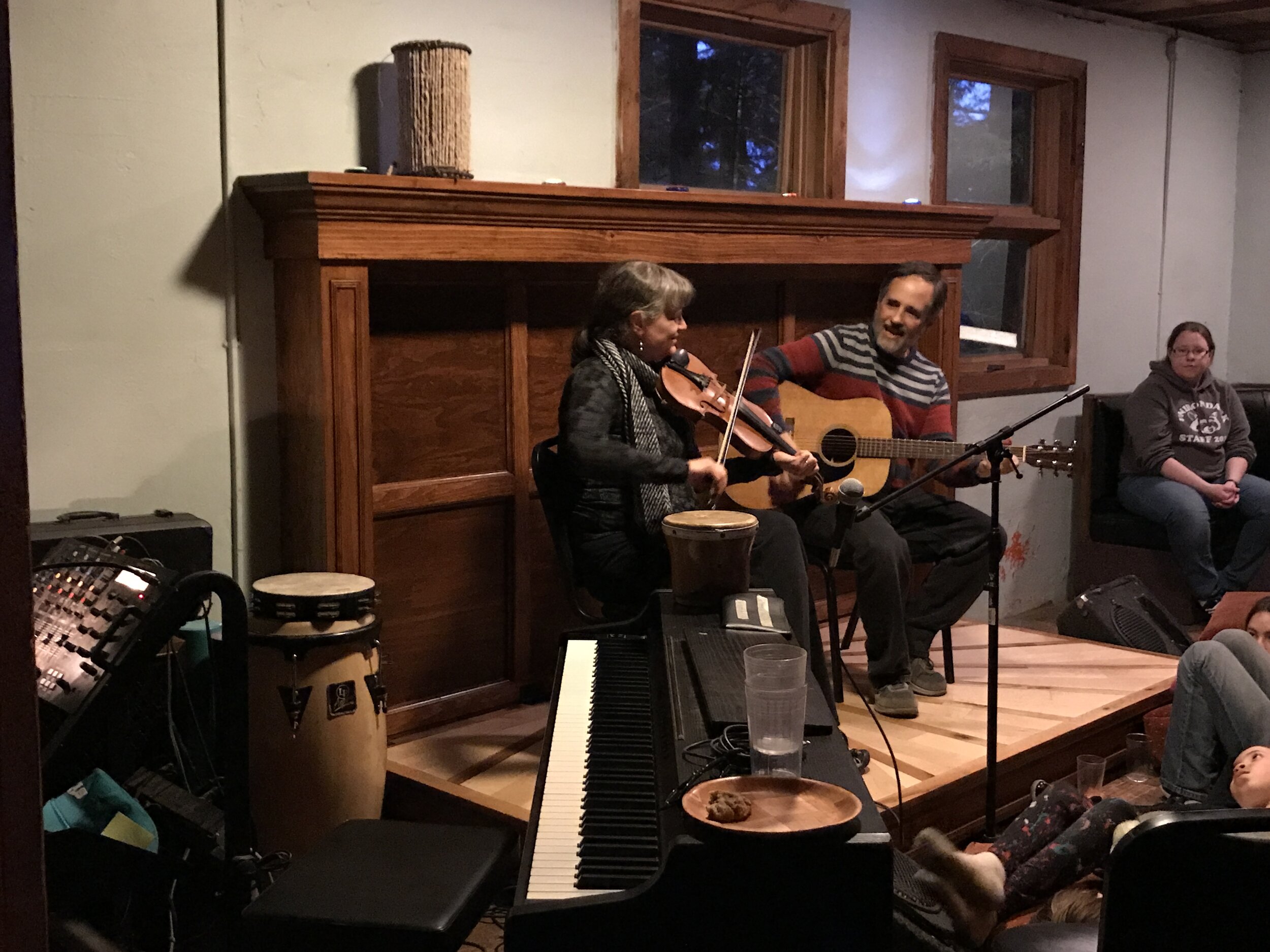 A woman and a man are performing music together on a small stage in a cozy indoor setting. The woman is playing a violin, and the man is playing an acoustic guitar and singing. There are musical instruments and equipment in front of them, including a piano, bongos, and a keyboard. Audience members are sitting and watching, with some visible in the foreground and background.