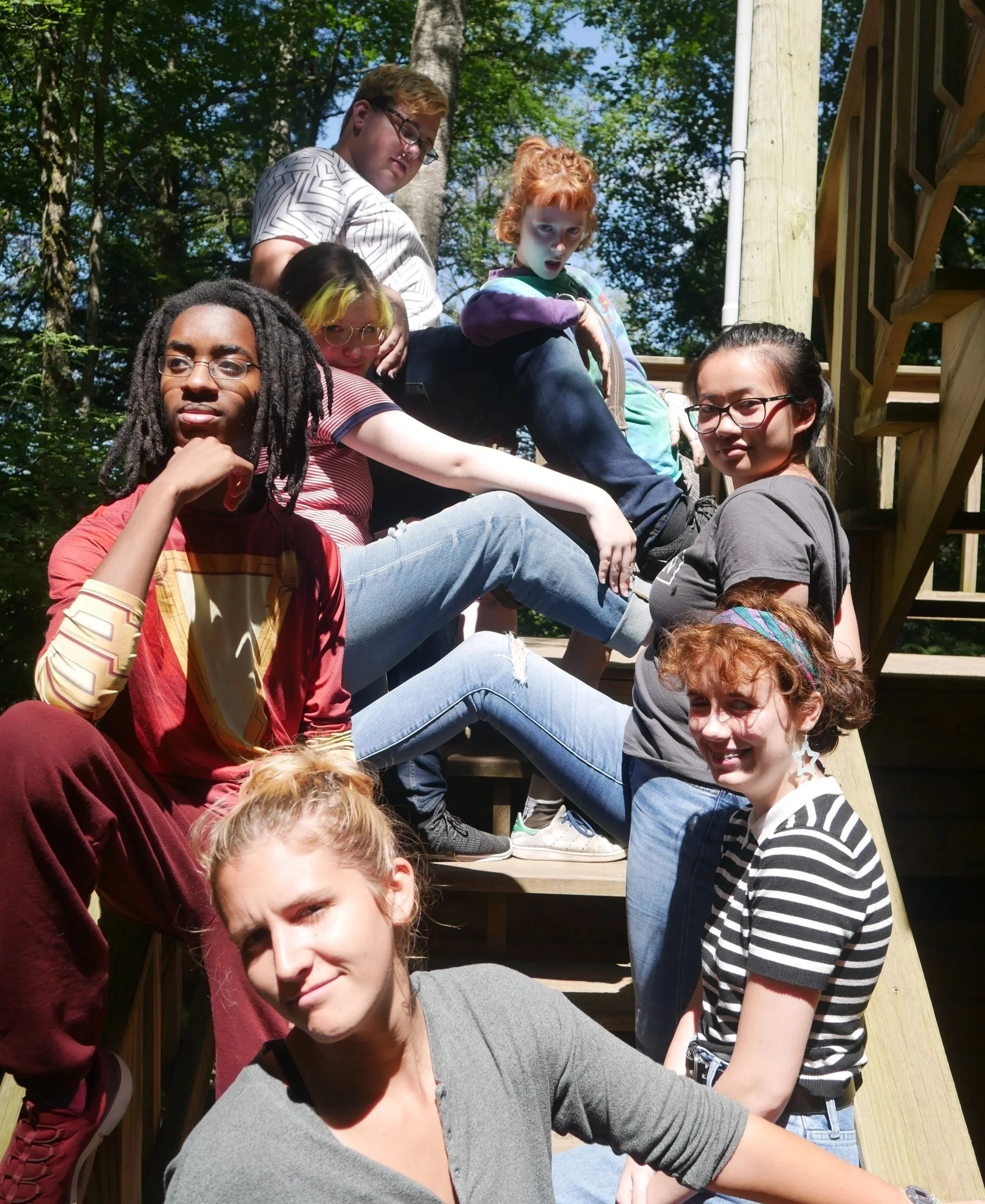 Group of nine young people sitting on a wooden staircase outdoors with trees in the background.