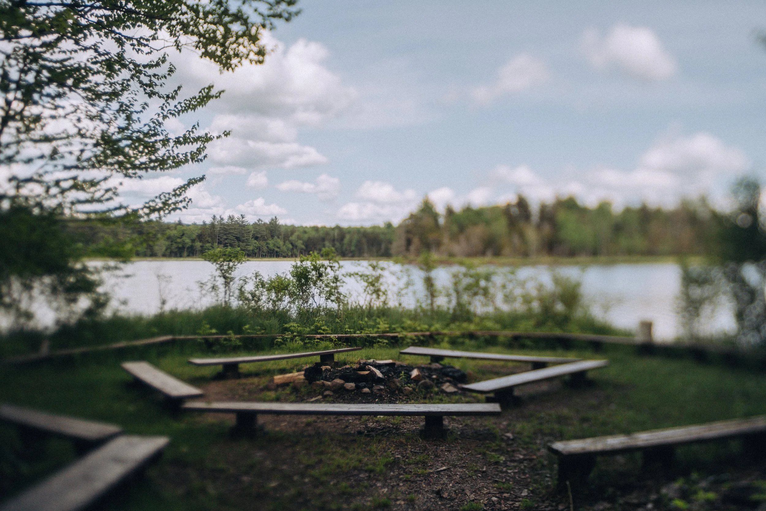 A lakeside camping area with a circular fire pit surrounded by wooden benches under a cloudy sky.