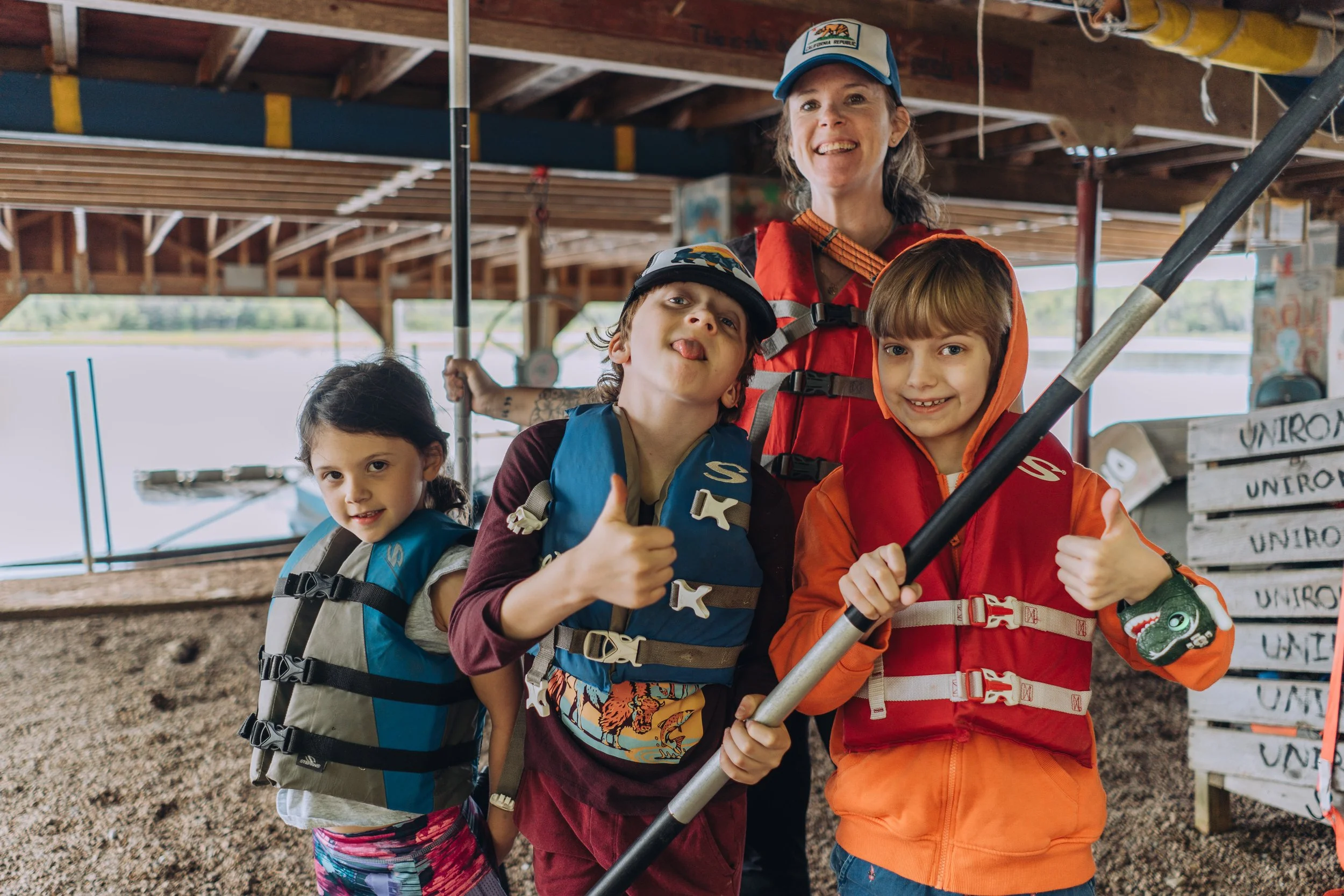 A group of four children and an adult woman on a boat dock, wearing life jackets and casual clothing. The woman is smiling and standing behind the children, who are posing with excitement, one of them giving a thumbs-up. The children are holding paddles, and the background shows a lake and wooden boat structures under a roof.