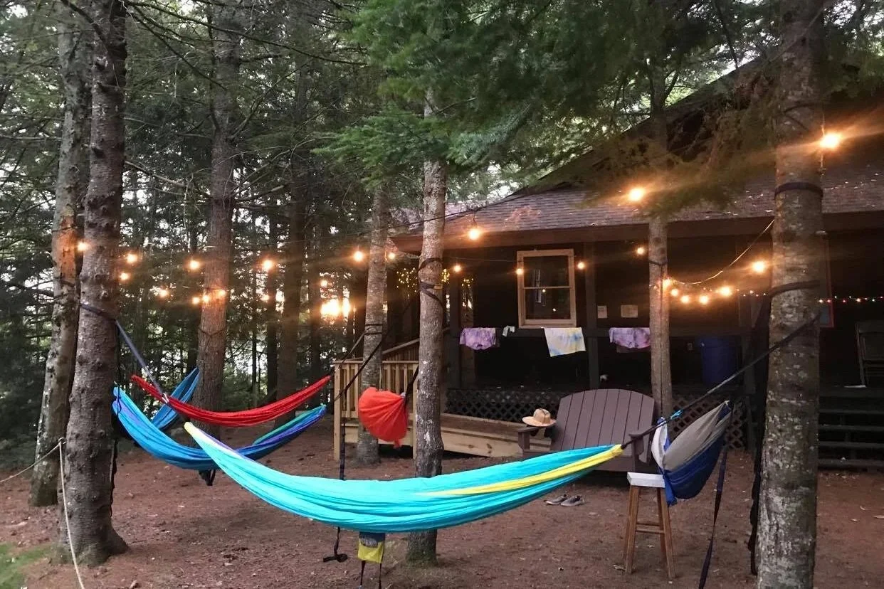 Two hammocks hung between trees with string lights overhead, in front of a cabin with towels hanging to dry.