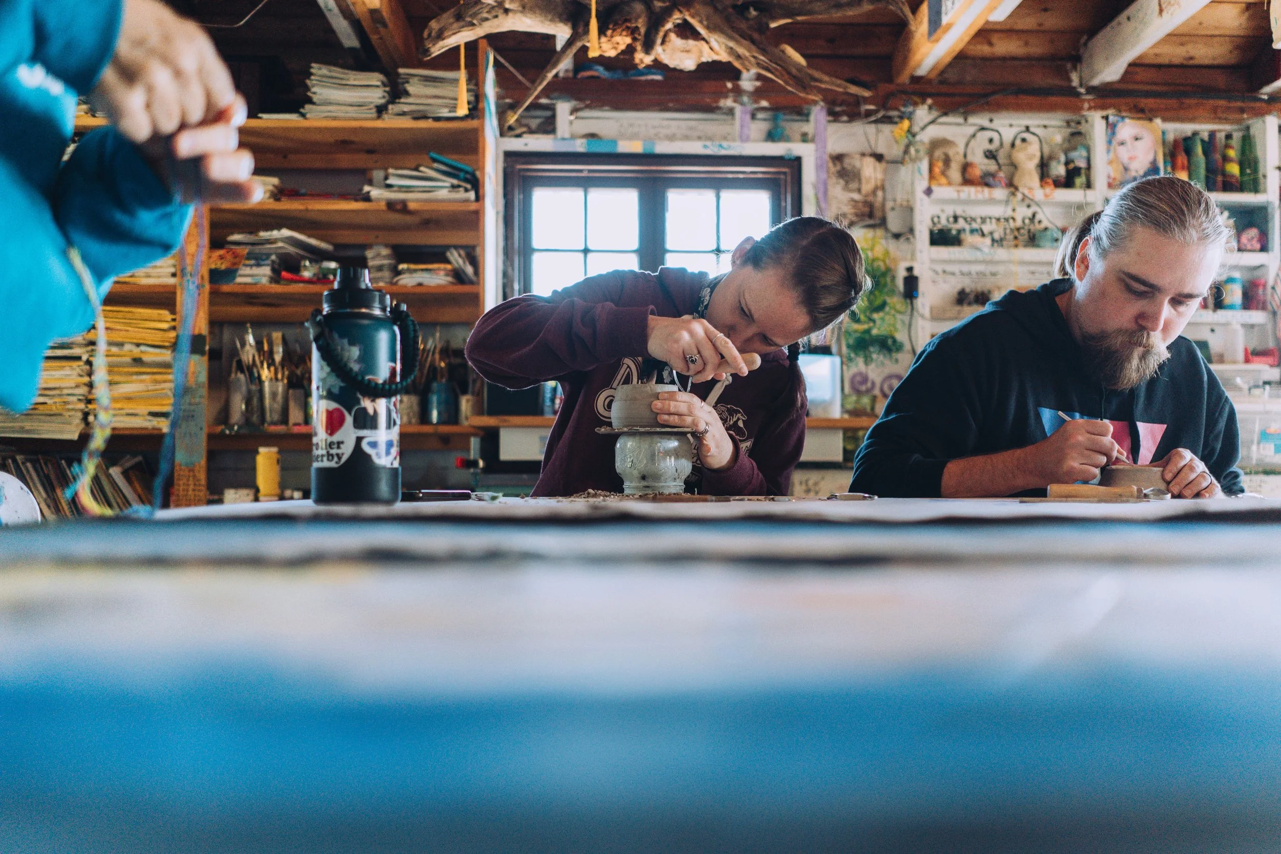 Two people working on pottery in a ceramic studio, with shelves of art supplies and a window in the background.