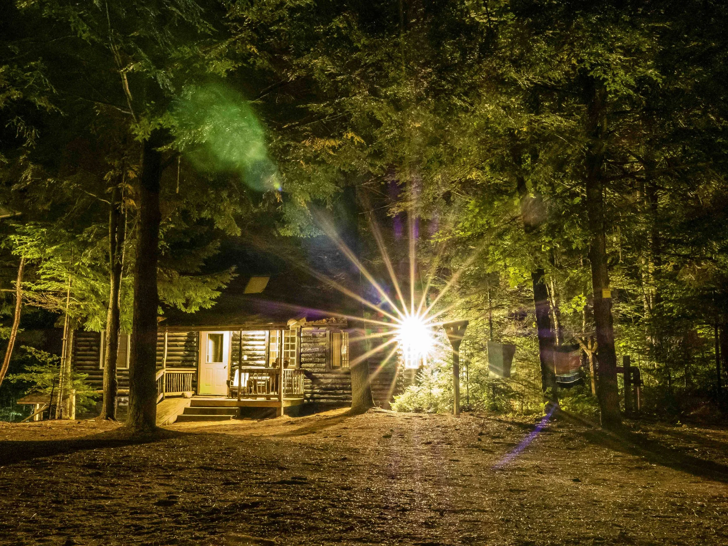 Nighttime scene of a wooden cabin in a forest with a bright light source shining directly behind it, illuminating trees and creating lens flare effects.