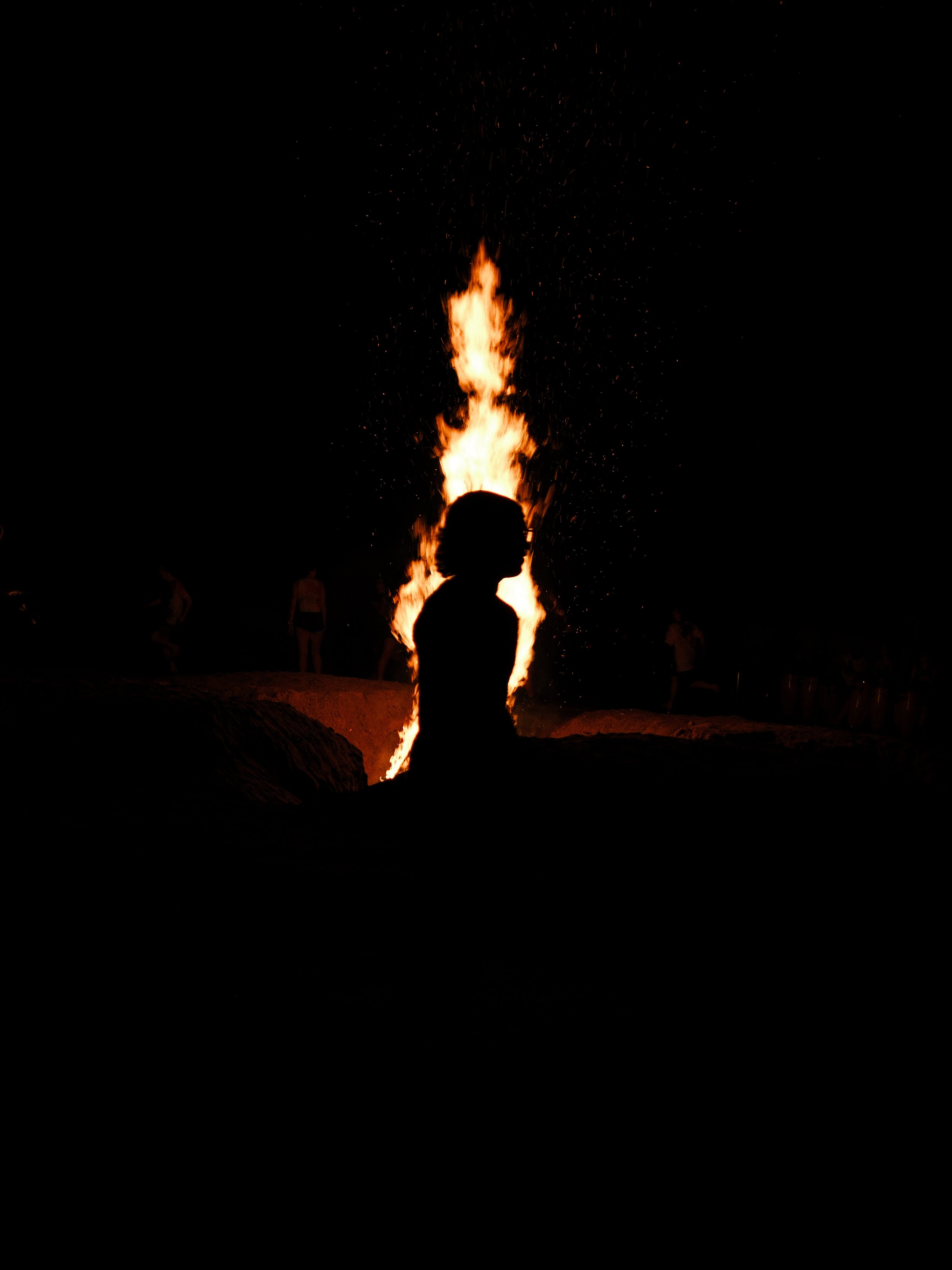 Silhouette of a person sitting near a large bonfire at night, with a dark background and other people in the distance.