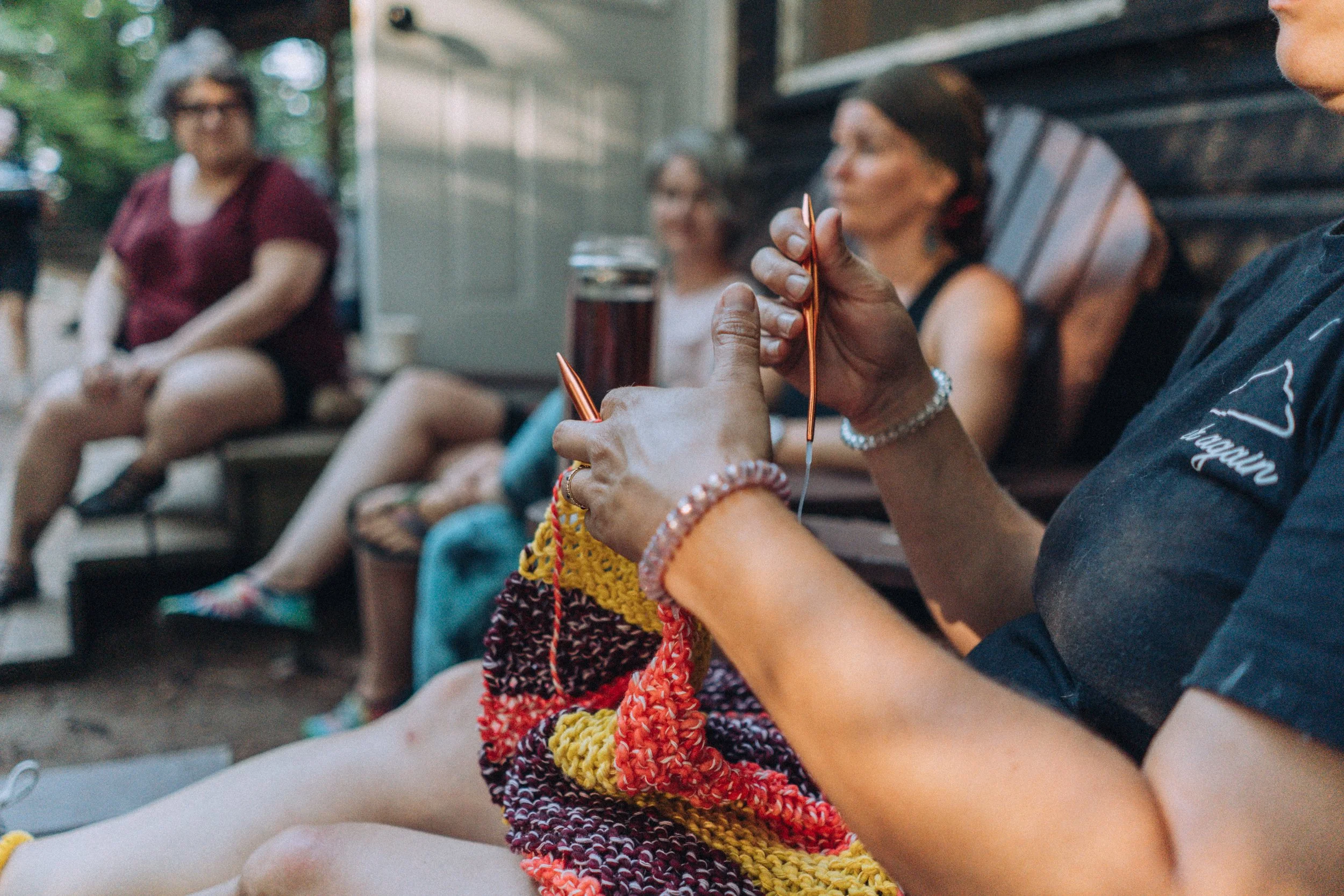 Person knitting colorful yarn while sitting outdoors with a group of women sitting on benches in the background.