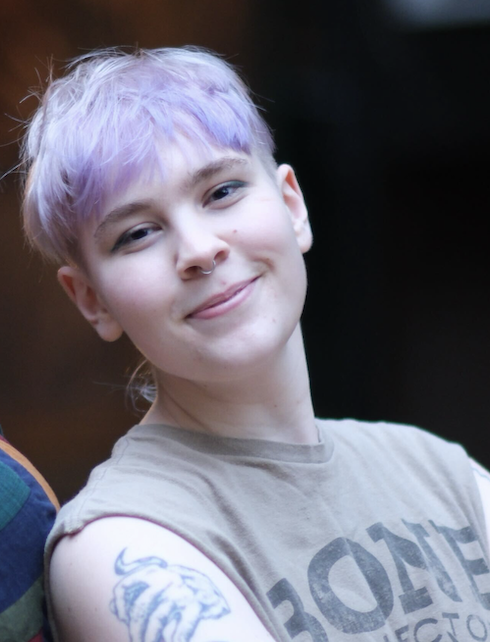 A person with teal-colored hair, wearing a brown Carhartt beanie and a dark shirt, sitting in an old, weathered white vehicle with a small black and white dog wearing a red bandana in their lap, both looking at the camera.