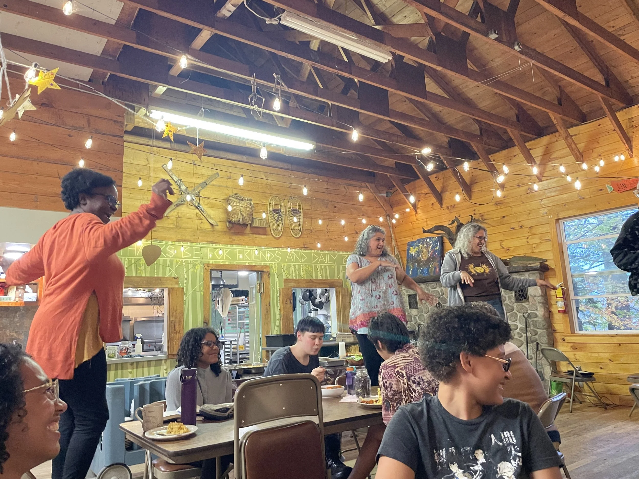 Group of people enjoying a social gathering in a cozy, rustic wooden room decorated with string lights, hanging stars, and other decor, some are standing and singing or dancing, while others are sitting at tables eating and chatting.