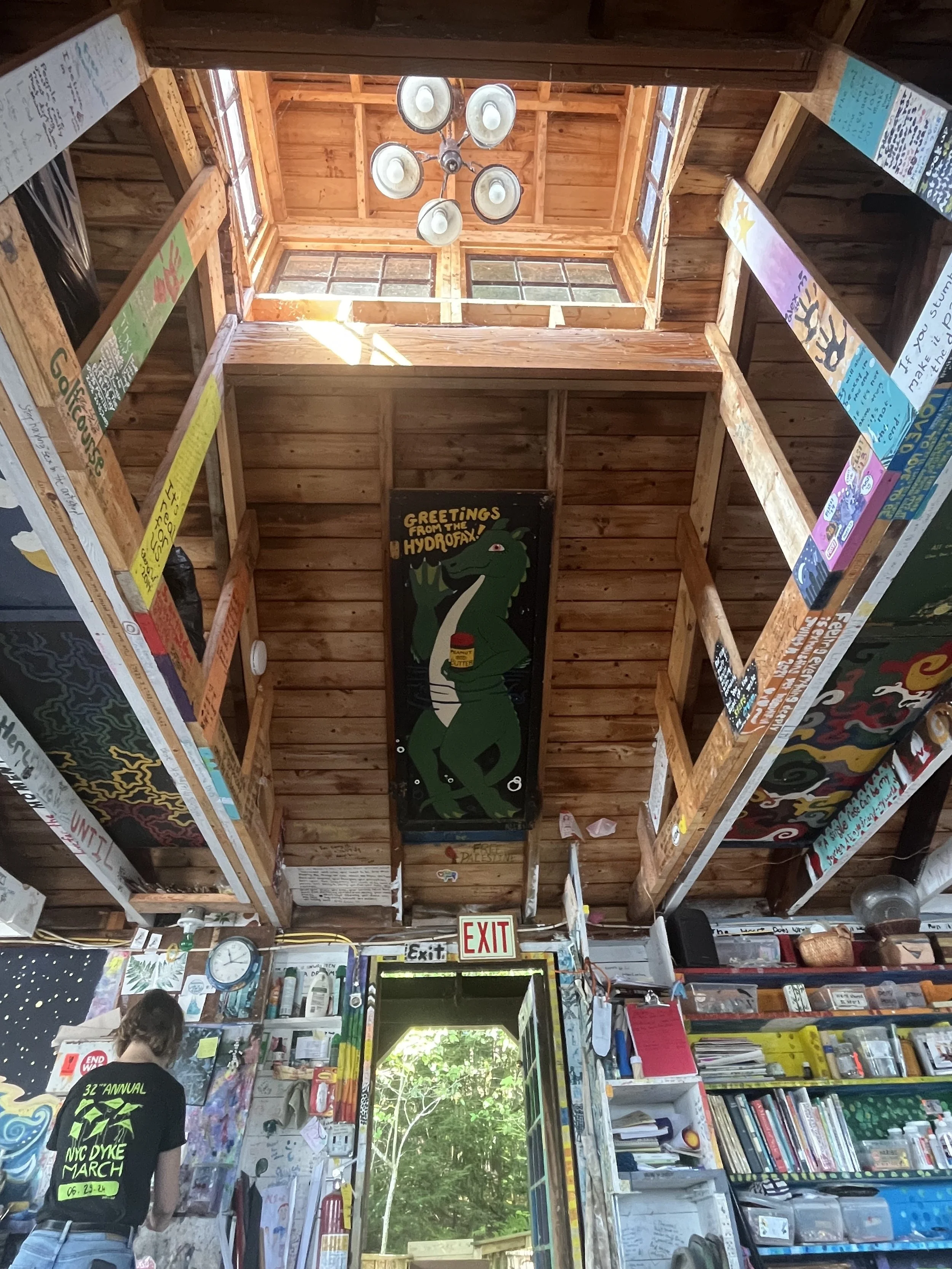 Interior view of a with wooden walls and a high ceiling, featuring a large window at the top letting in natural light, a chandelier, colorful handwritten signs and artwork on the walls, a prominent sign with a cartoo saying 'Greetings from the Hydrofax!', and shelves filled with books and supplies.