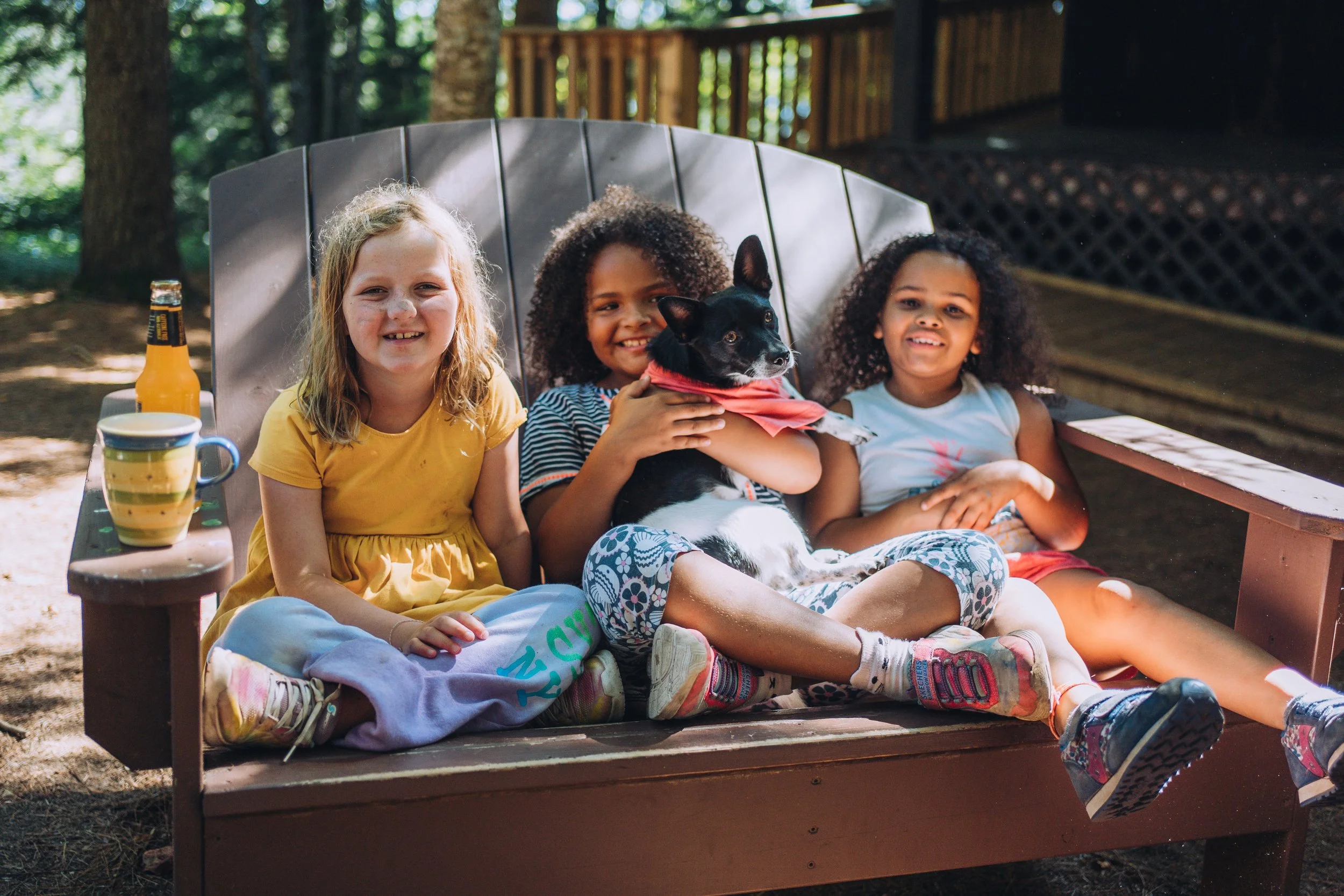 Three girls sitting on a bench outdoors with a small black dog, two bottles, and a mug on the side.