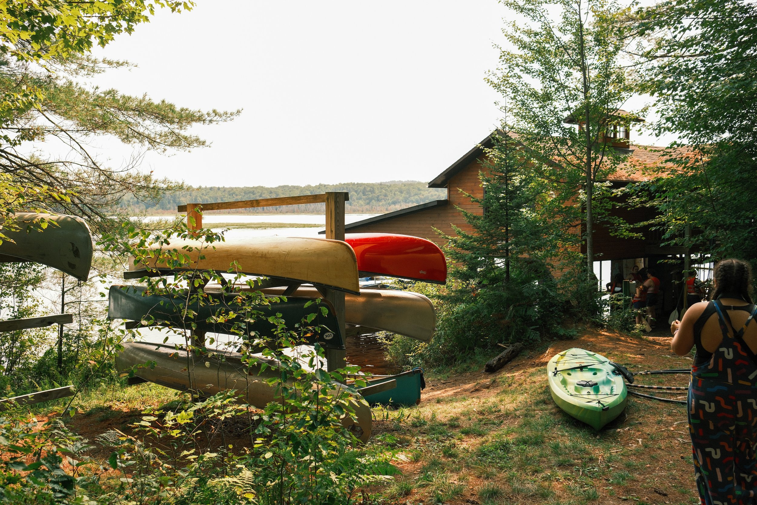 Kayaks stacked on a rack near a lakeside at a summer camp, with a green paddleboard and a person with braided hair and colorful pants in the foreground.