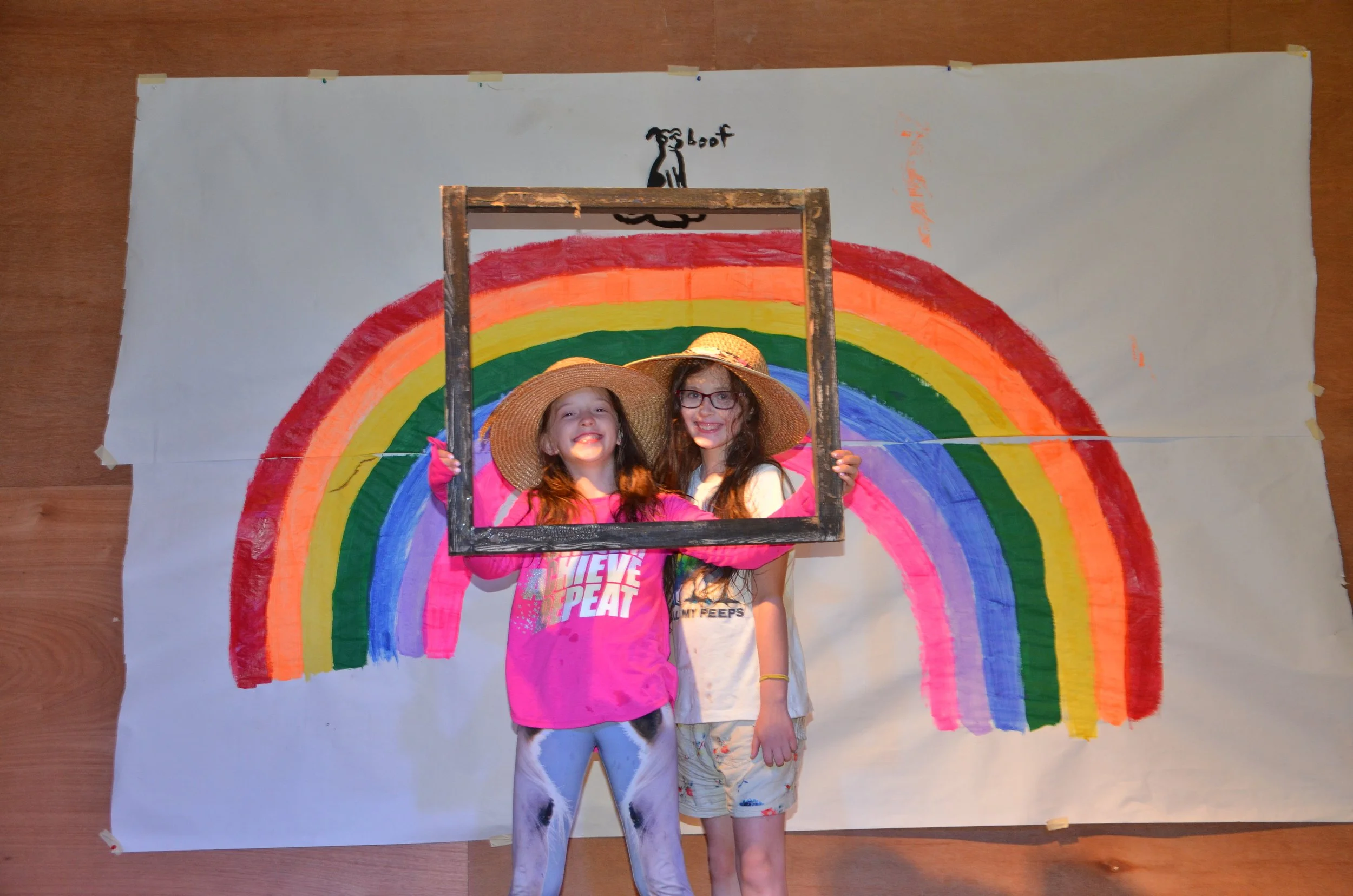 Two girls wearing straw hats pose behind a wooden frame in front of a large rainbow drawing on a white paper backdrop. One girl is wearing a pink shirt with 'Achieve and Repeat' printed on it, and the other is in a white shirt and shorts. The rainbow is painted with bands of red, orange, yellow, green, blue, and purple, and a small dog drawing with the word 'woof' is above the rainbow.