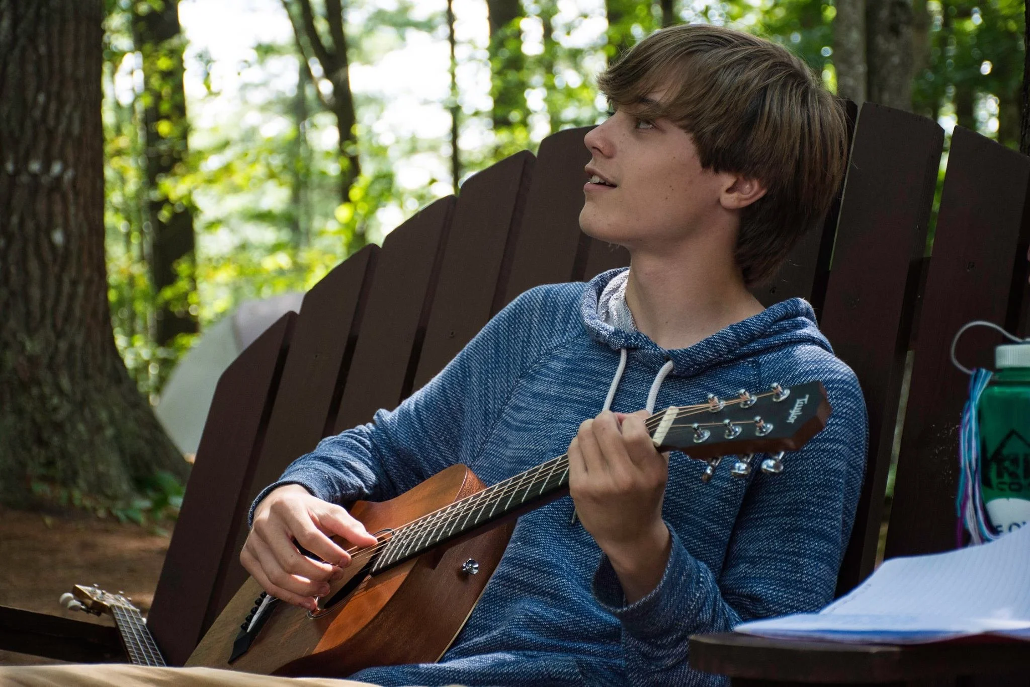 A young man with brown hair sitting on a wooden bench outdoors, holding an acoustic guitar and looking to the side, surrounded by trees and greenery.