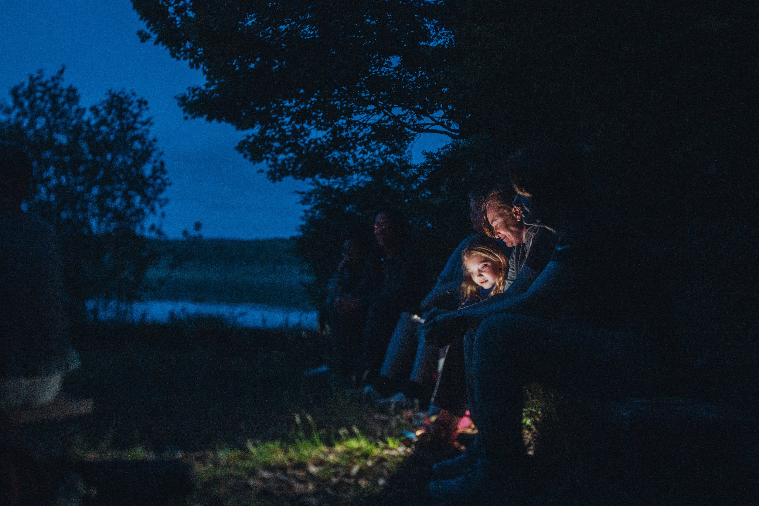 A group of people sitting outdoors in nature during the evening, gathered around a small campfire, with a body of water and trees in the background.