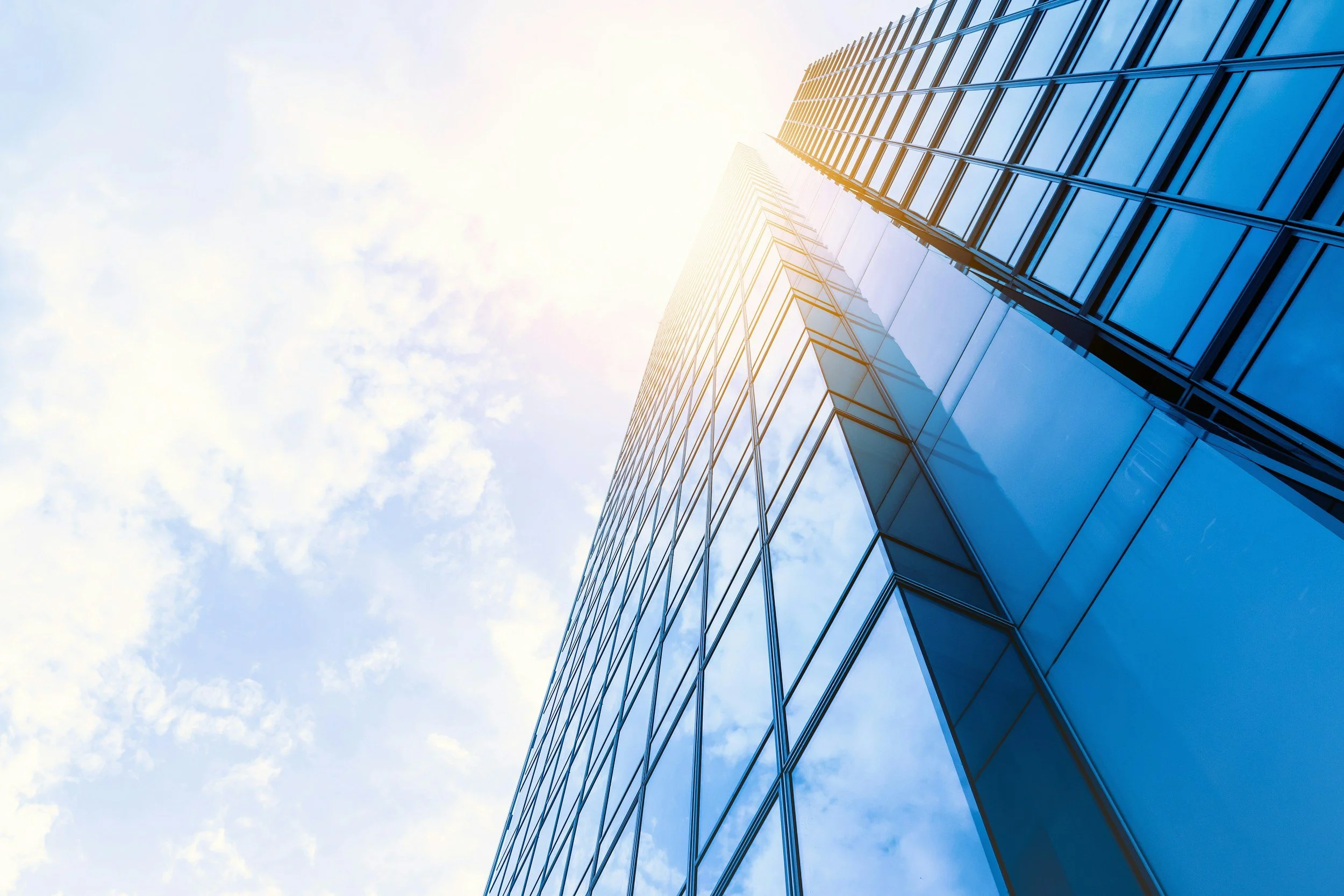 Looking up at a modern glass skyscraper against a partly cloudy sky with the sun shining brightly.