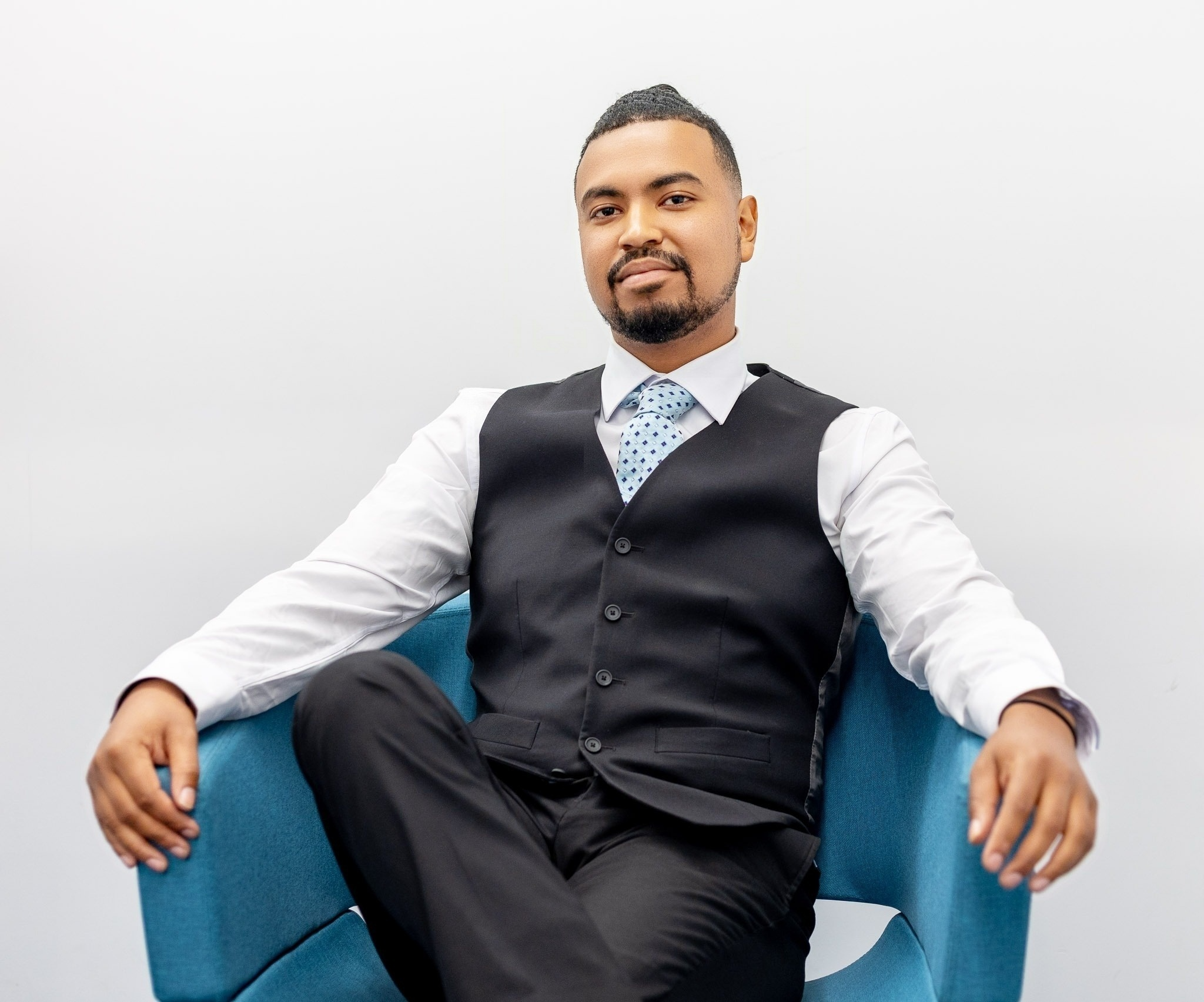 Man sitting on a blue chair, wearing a white shirt, a black vest, and a blue patterned tie, against a plain white background.