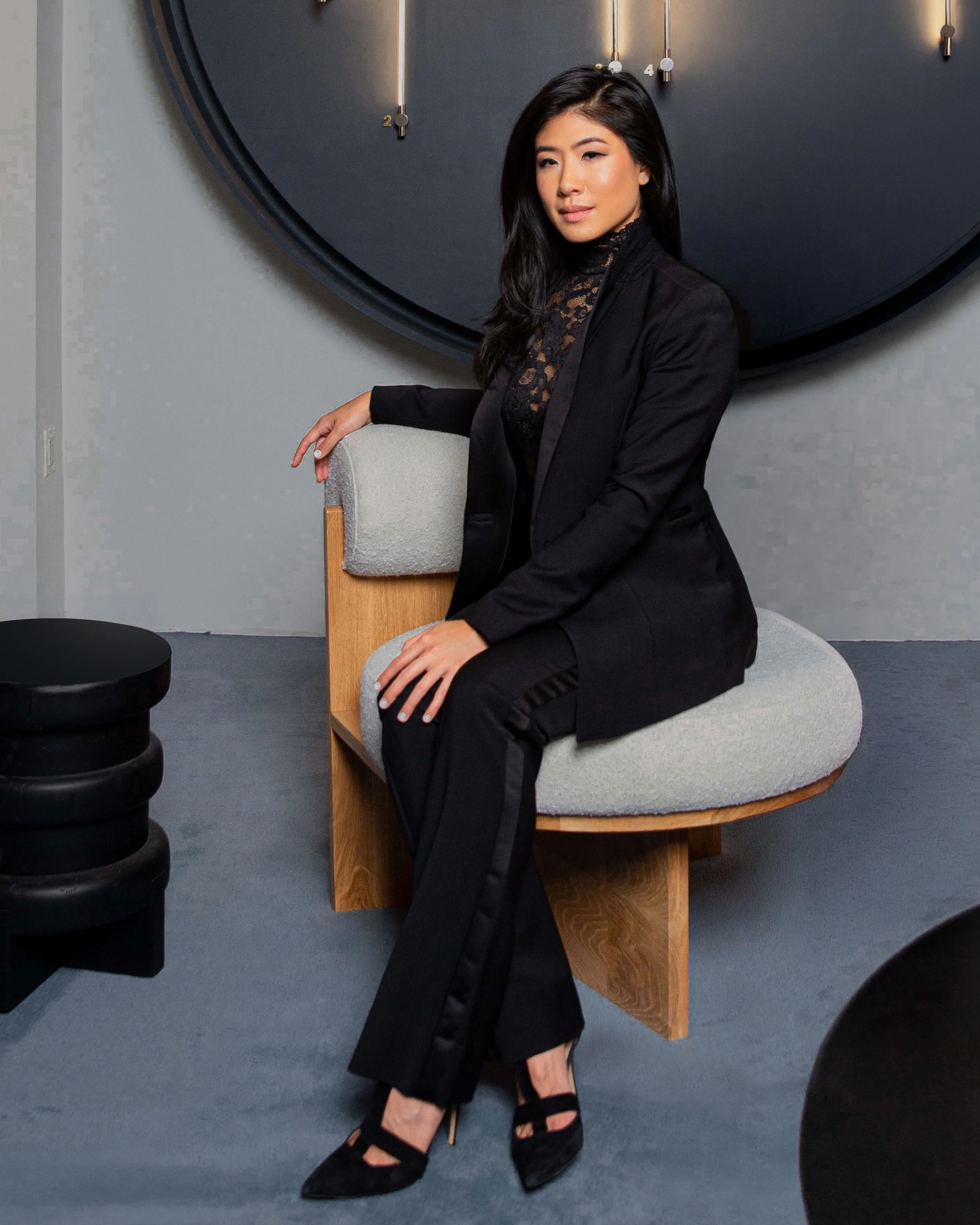 A woman with long dark hair wearing a black blazer and black pants sits on a modern wooden and cushioned chair in a contemporary room with a large dark wall clock in the background and a small black table in the foreground.