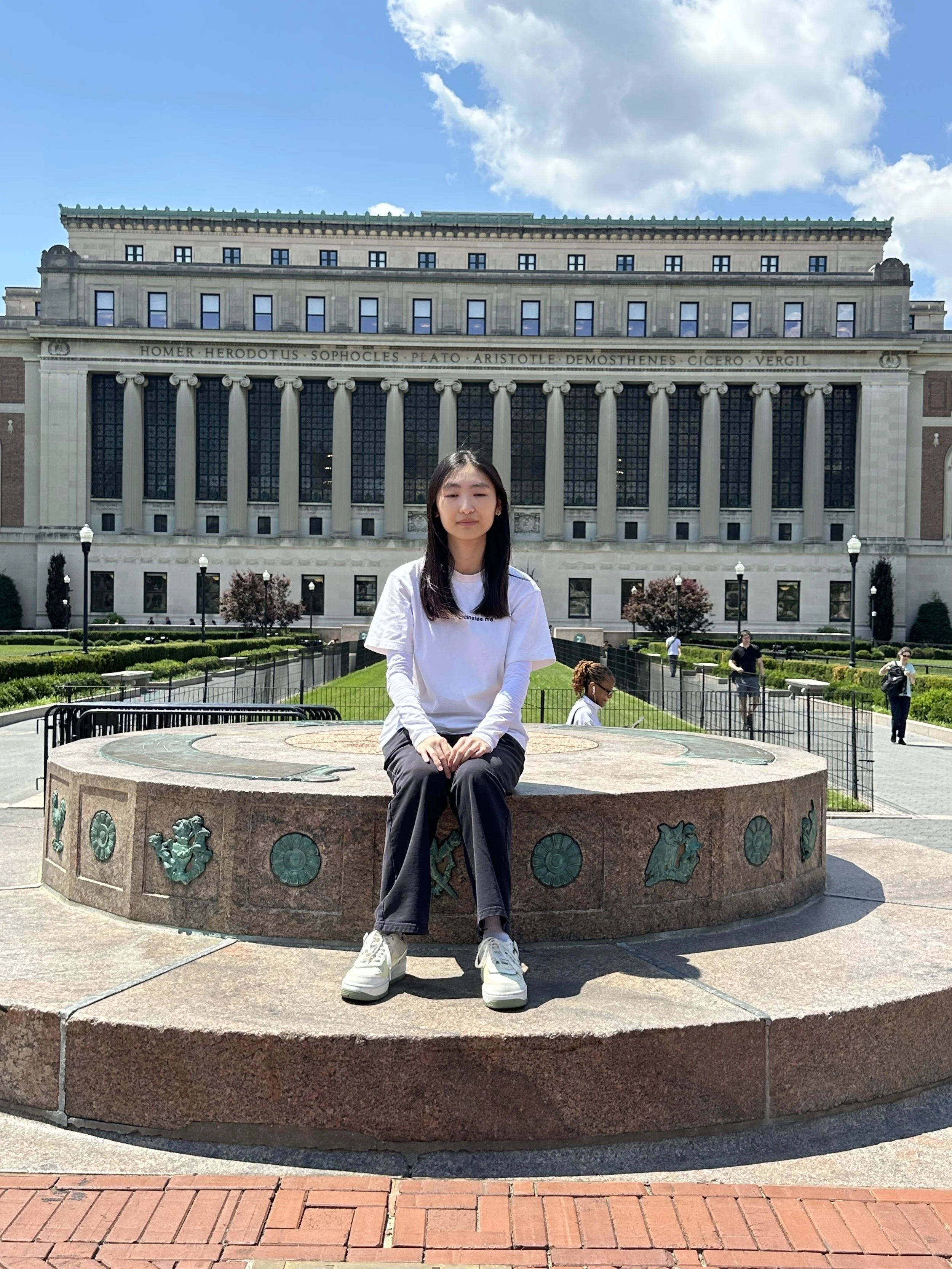 A young woman with long dark hair and light skin, wearing a white long-sleeve shirt and black pants, sits on a stone fountain in front of a large classical-style building with columns and an inscribed frieze. The building has a clear blue sky with some clouds above.