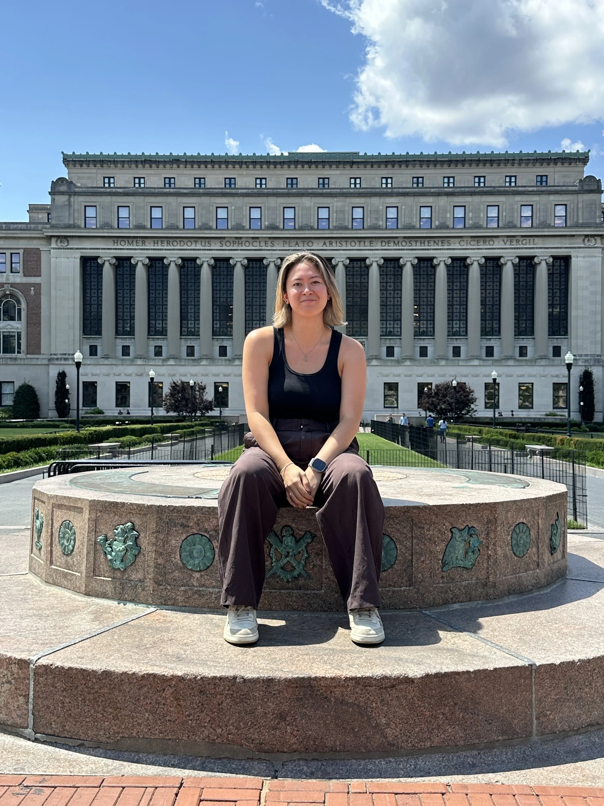 A woman sitting on a decorative stone bench in front of a historic building with columns and books engraved on the top. The sky is blue with some clouds.