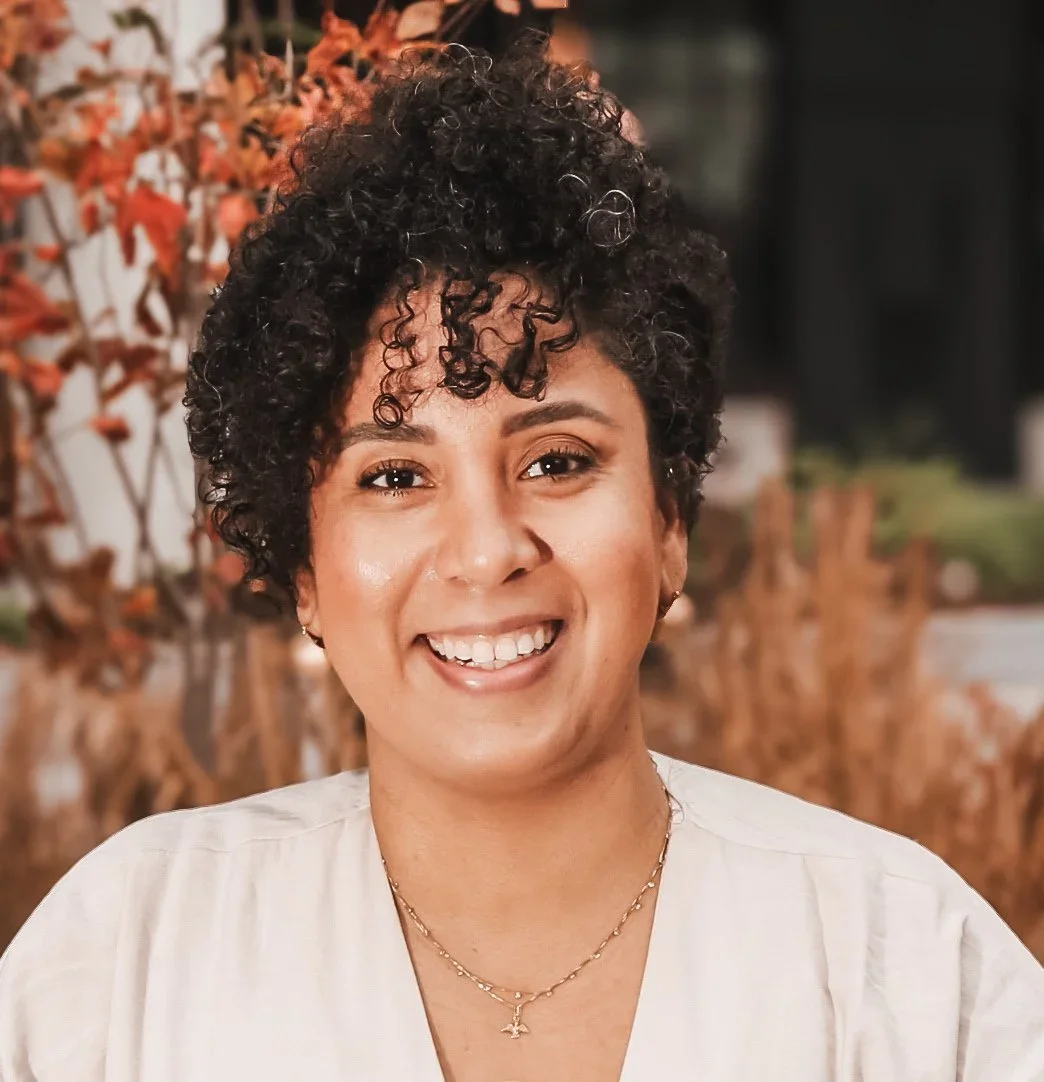 Close-up of a smiling woman with short, curly black hair, wearing a white top and a delicate gold necklace, outdoors with autumn-colored foliage in the background.