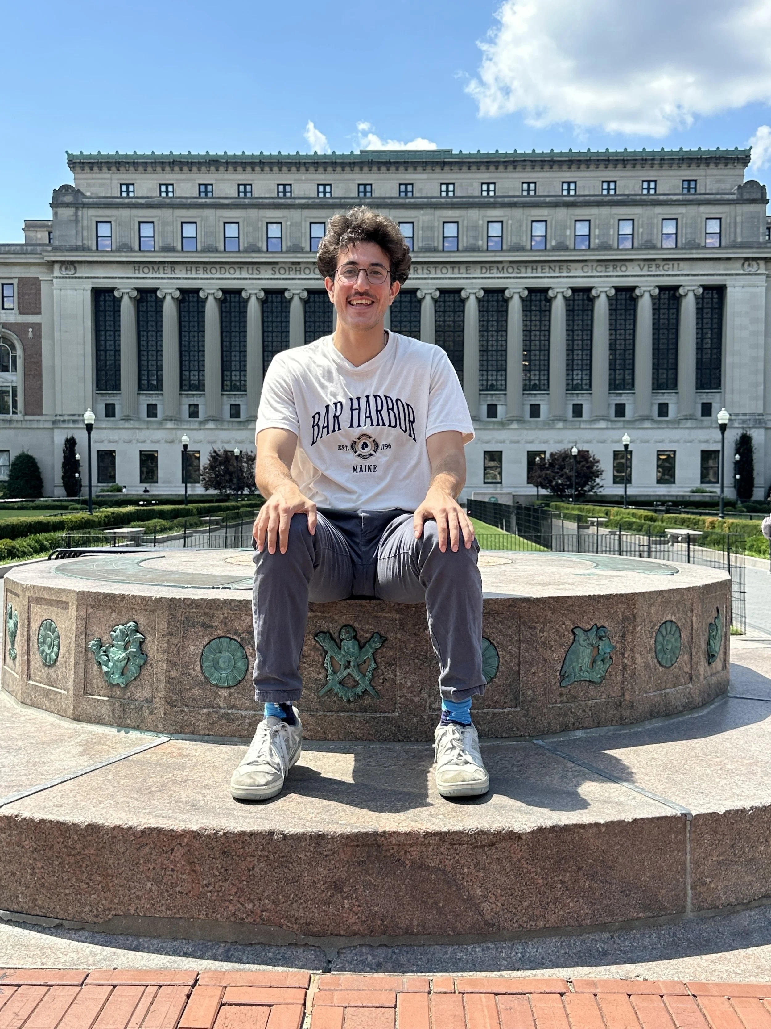 A young man sitting on a decorative stone fountain in front of a large, classical-style building with tall columns, under a partly cloudy sky.