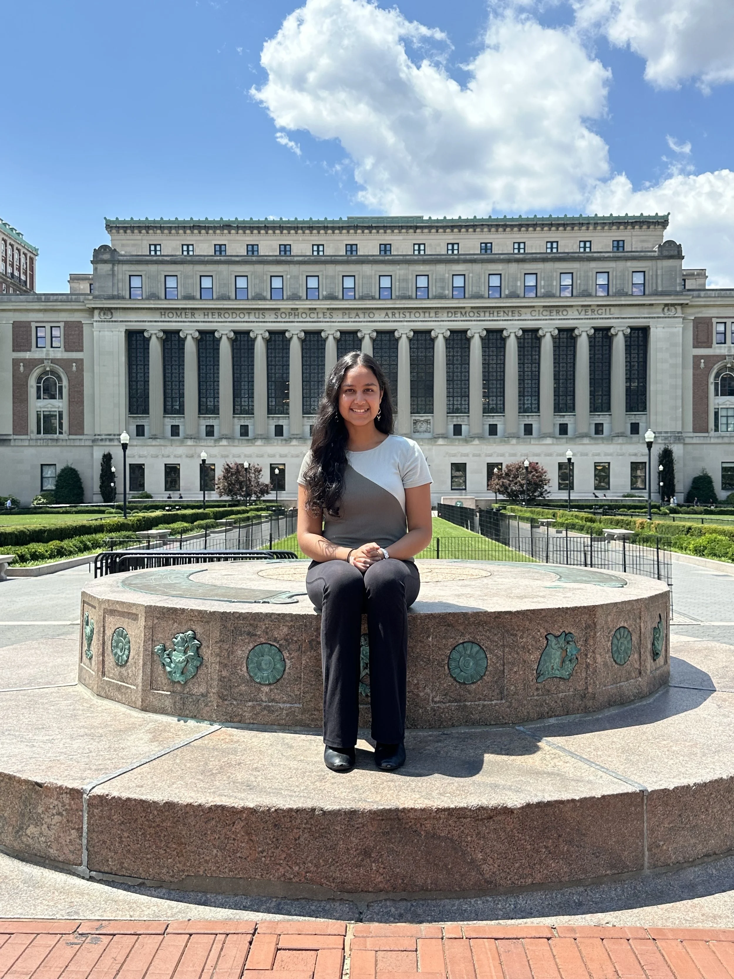 A woman sitting on a circular stone bench with decorative metal plaques, in front of a large, historic building with columns and inscribed names, under a partly cloudy sky.