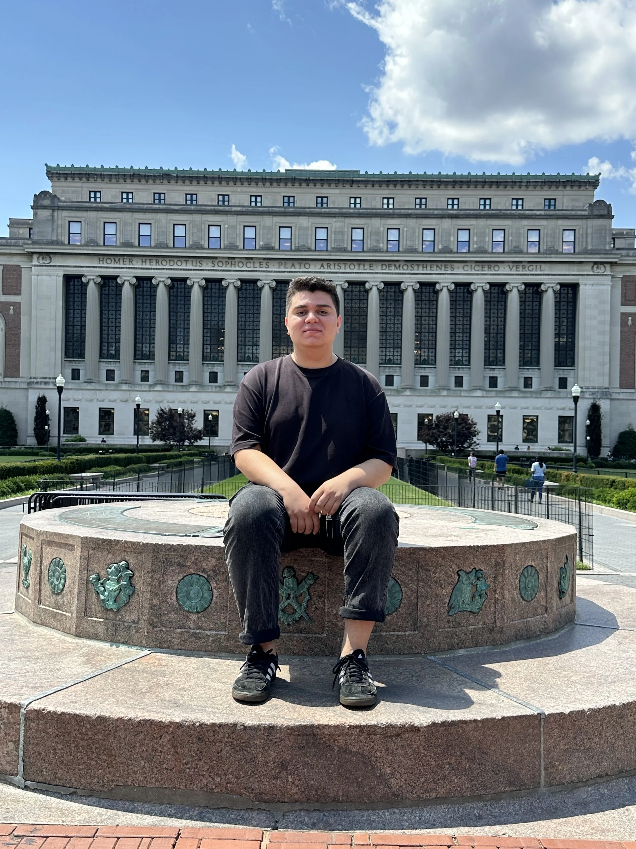 A young man sitting on a circular stone bench in front of a large historic building with columns and inscriptions, on a sunny day with blue sky and scattered clouds.