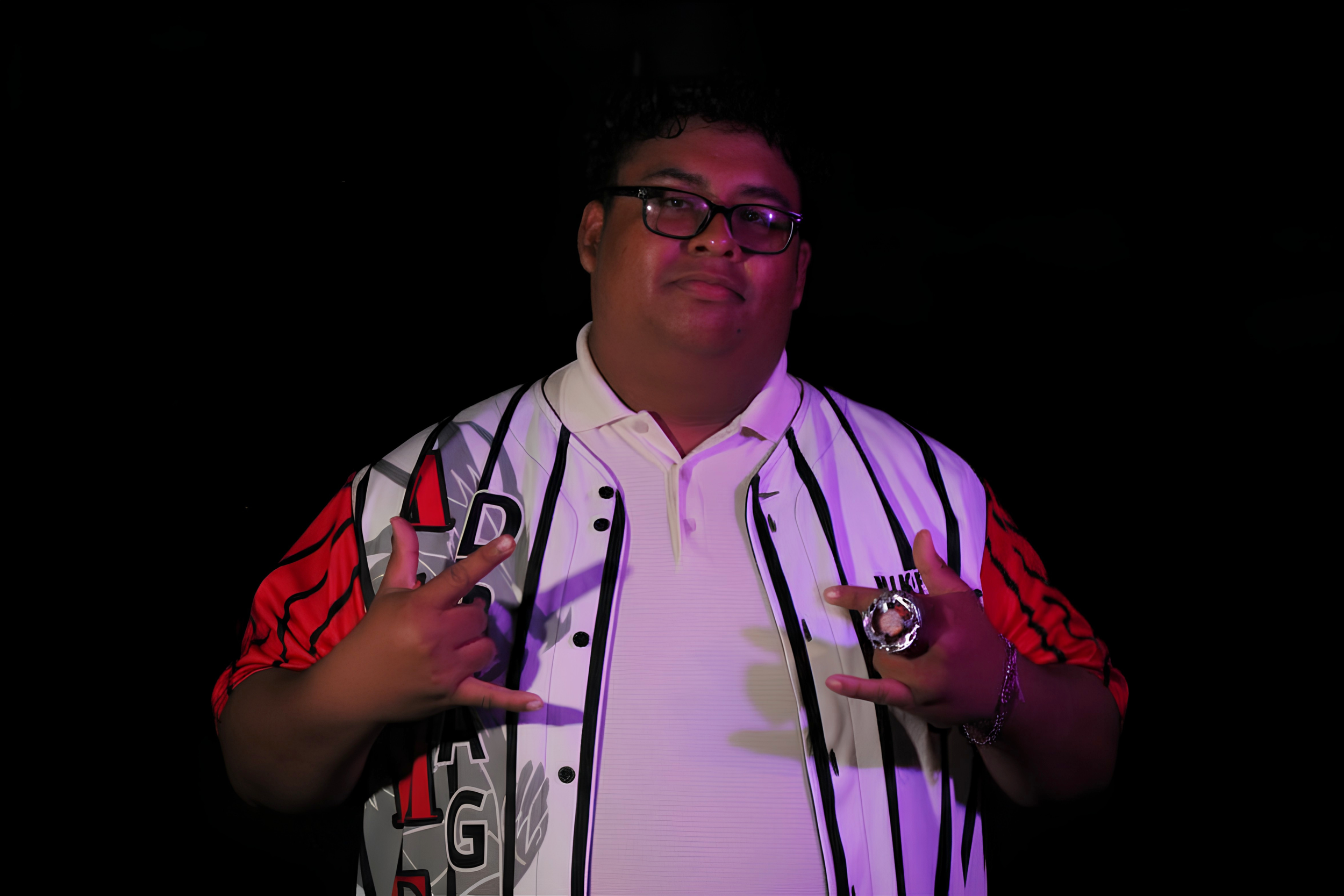 A young man with glasses wearing a white shirt and an open jacket featuring black, red, and gray graphics, holding up both hands in a rock and roll gesture against a dark background.