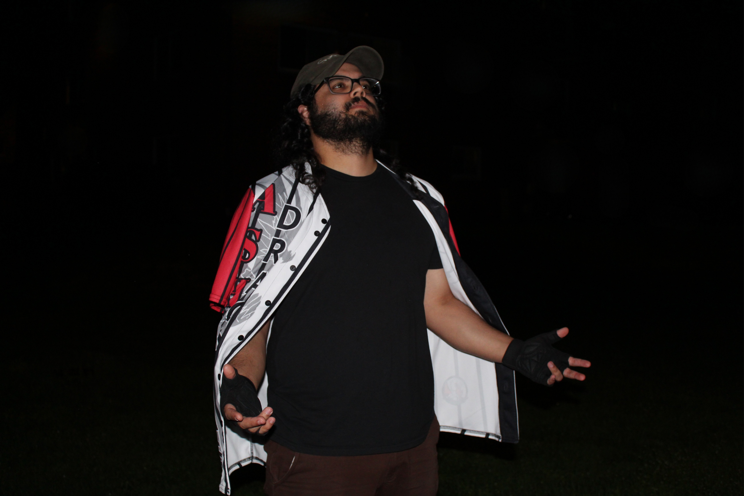 A man with long curly hair, beard, and glasses standing outdoors at night with hands gesturing outward, wearing a gray cap, a black T-shirt, and a white jacket with red, black, and white graphics.